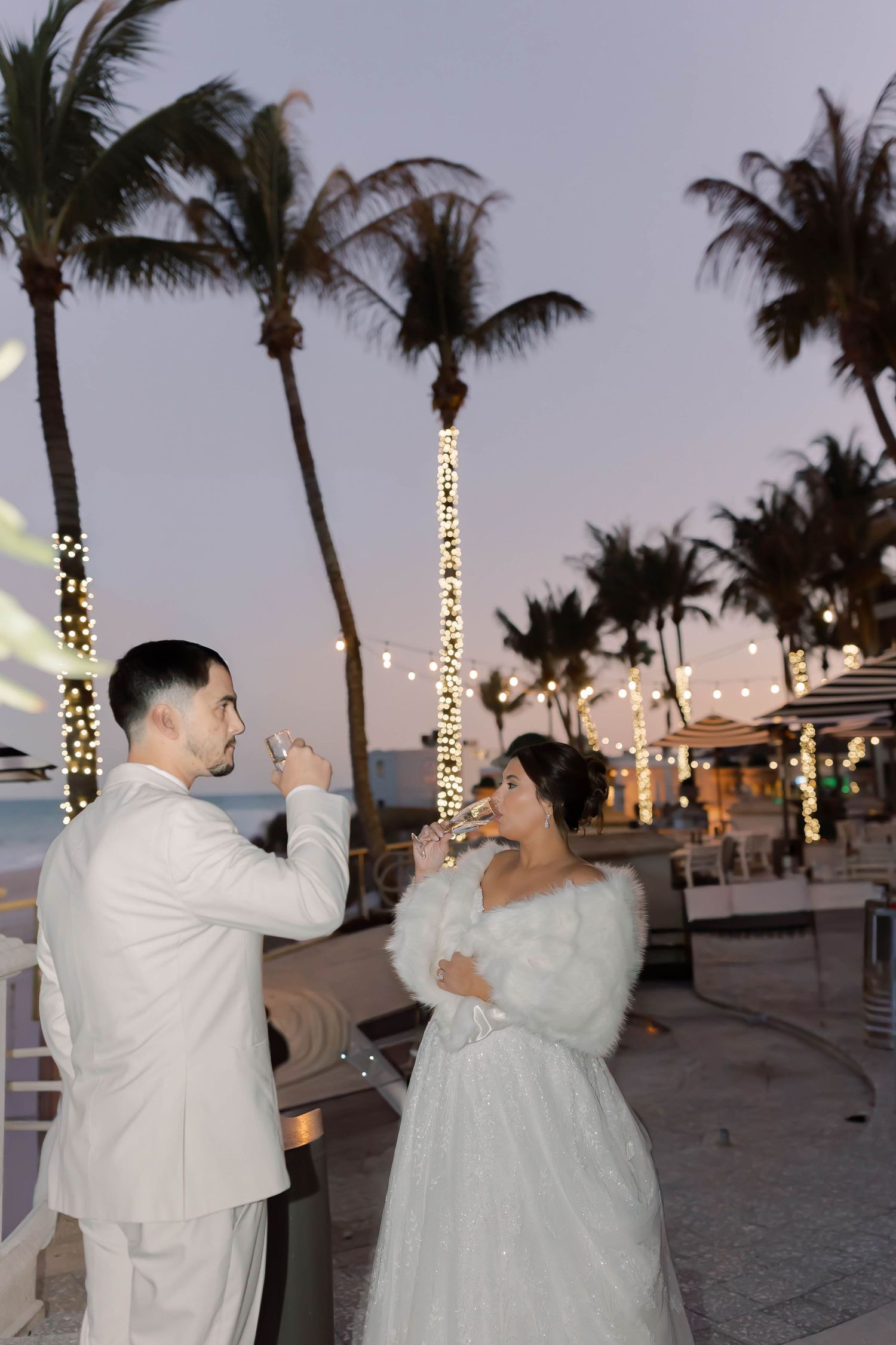 Bride and groom toasting with champagne at a tropical beach wedding venue at dusk with palm trees and string lights.