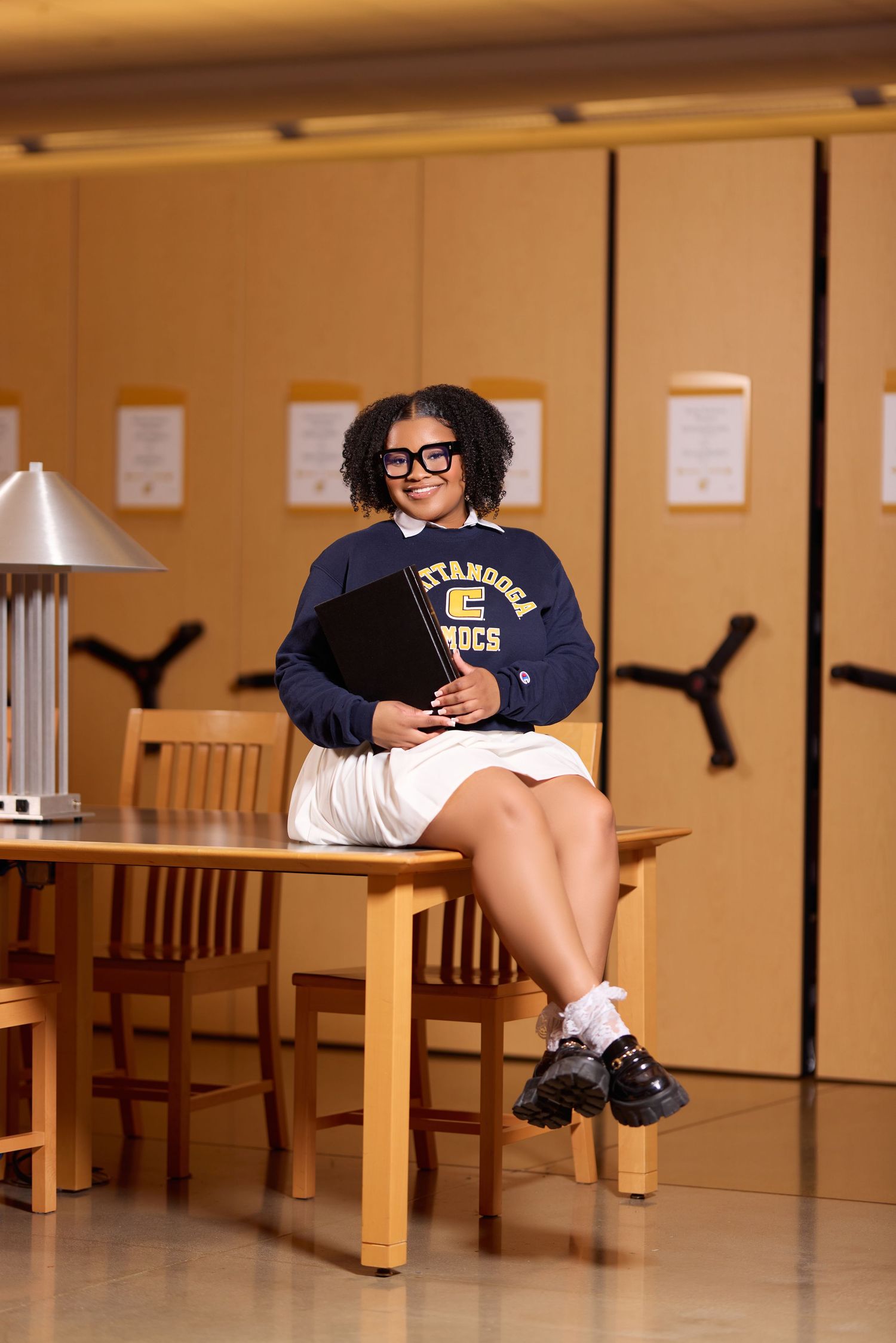 College student in navy sweater poses in study area with wooden furniture.