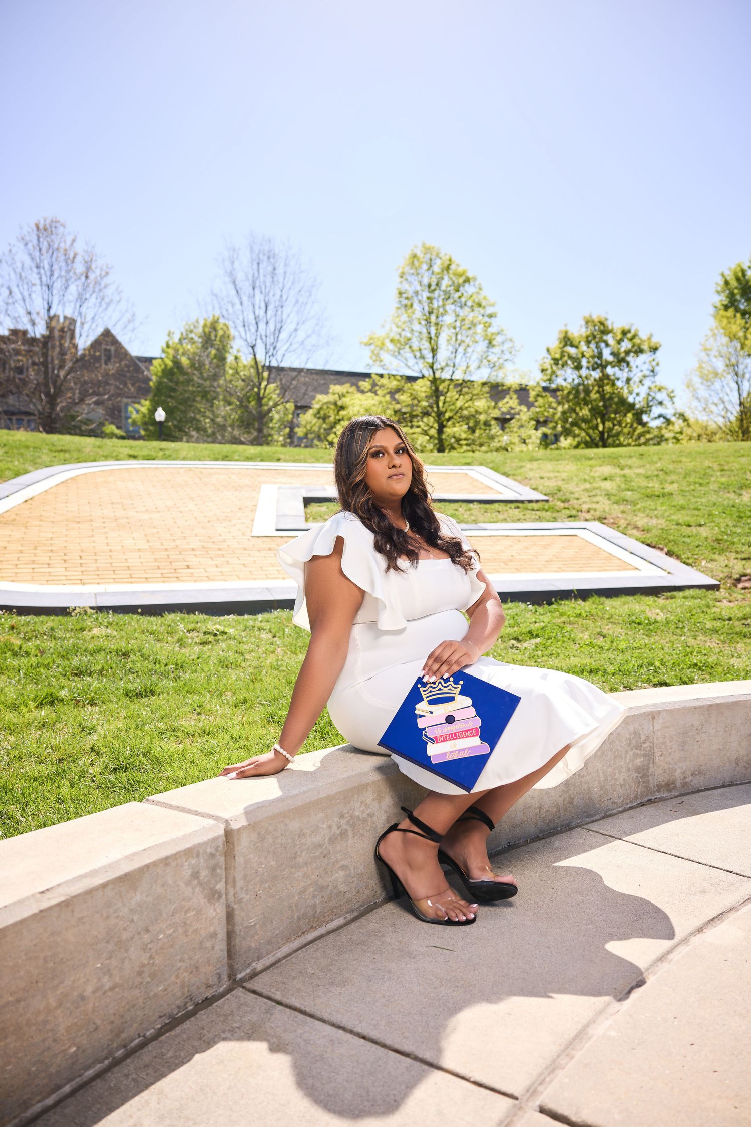 Graduate sits casually on concrete steps wearing graduation attire against campus backdrop.