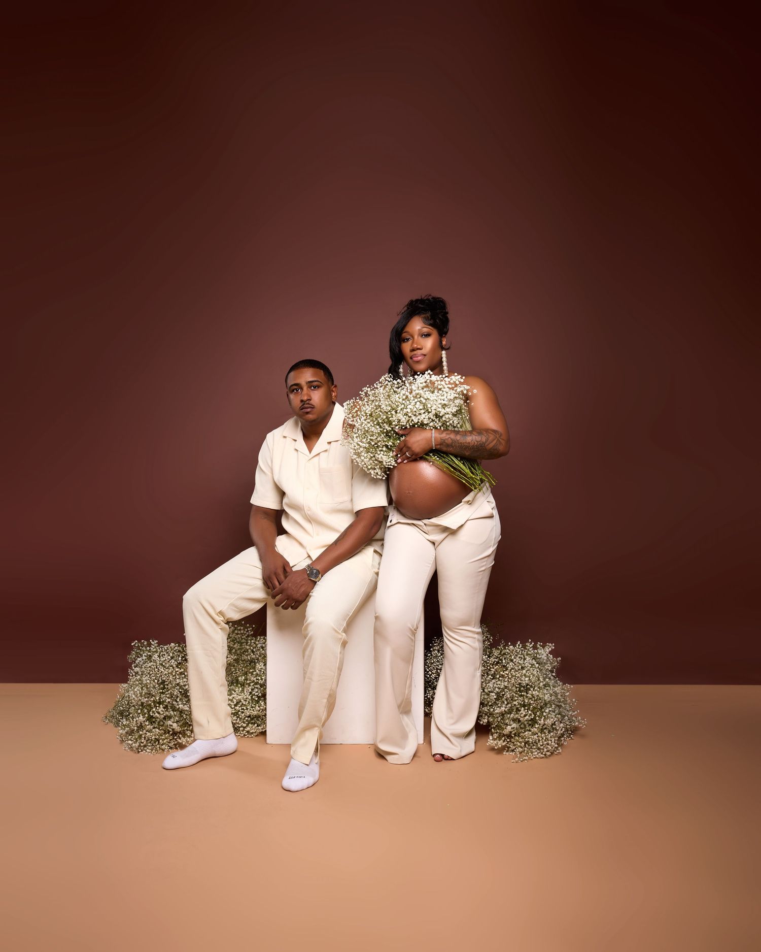 Artistic maternity photos with couple in white outfits against brown backdrop with baby's breath flowers.