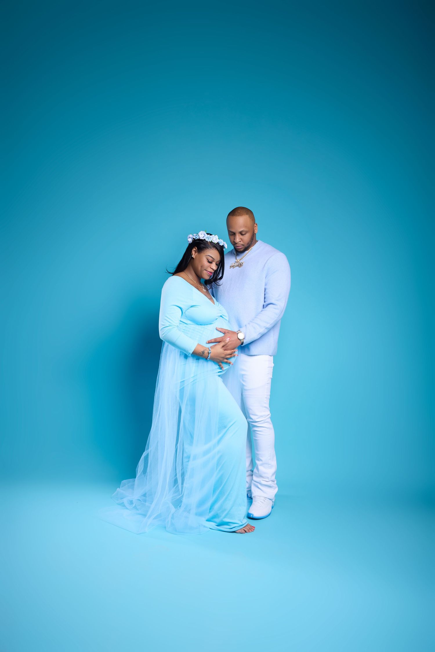 A pregnant woman in a blue gown with a man in white, both gazing lovingly at her baby bump on a blue background.