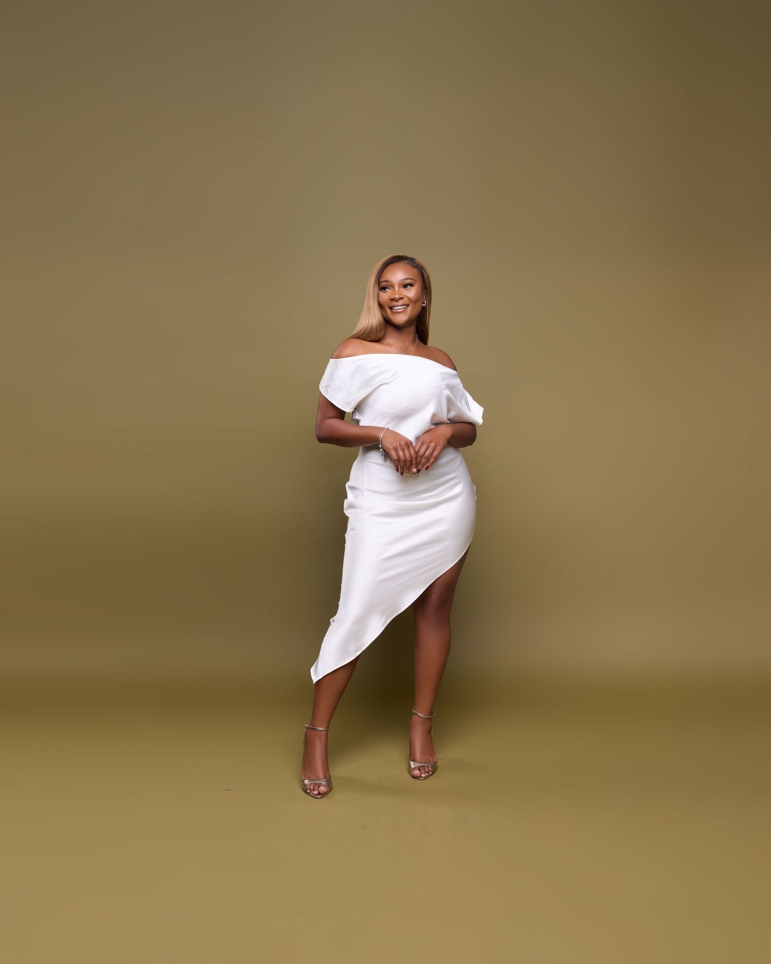 Model in white dress poses elegantly on chair against neutral backdrop in studio photoshoot.