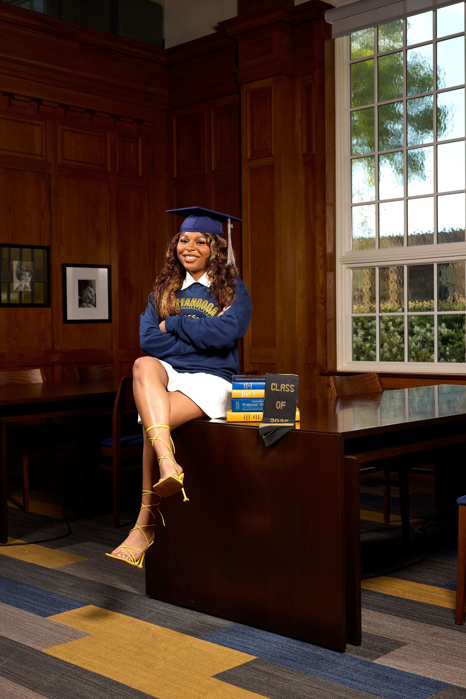 Graduate poses with books in wood-paneled study room with large windows.
