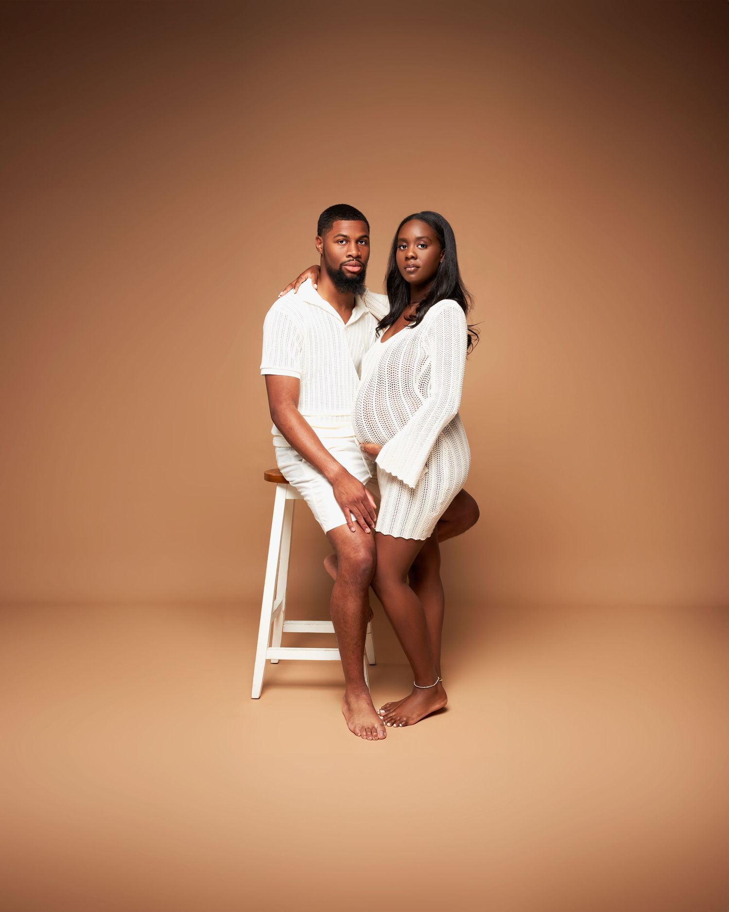 Intimate studio portrait of a couple in white outfits posing together against a brown backdrop.