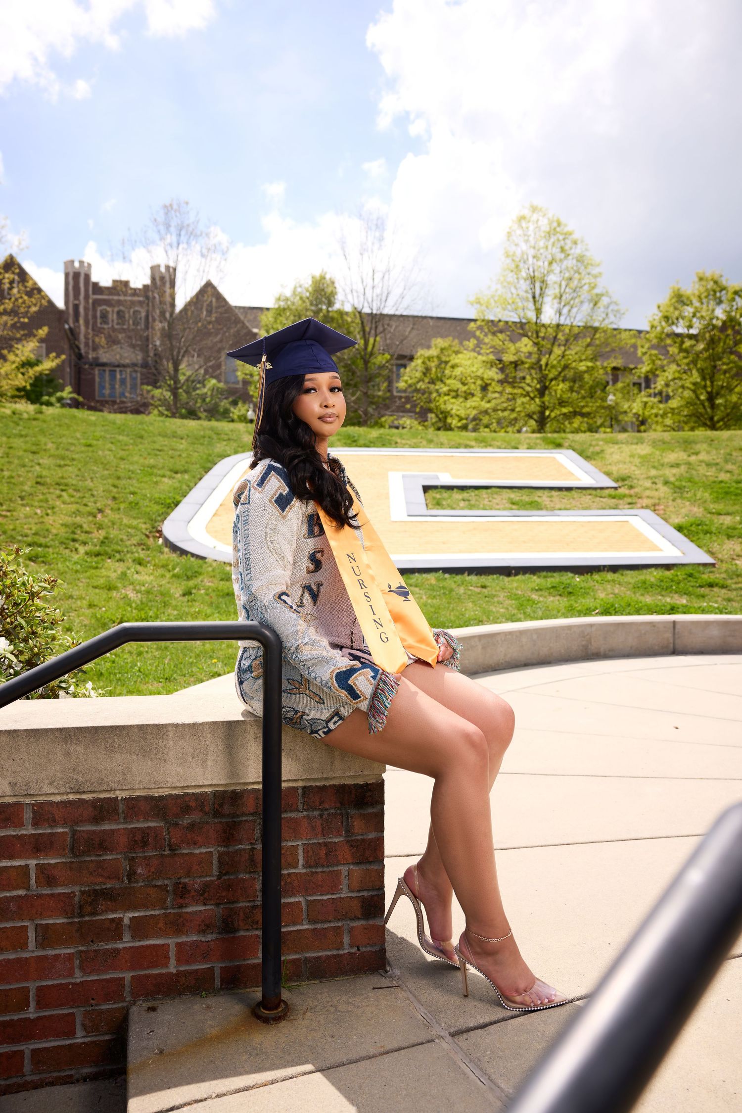 Graduate in cap and gown poses on brick wall with campus buildings visible in background.