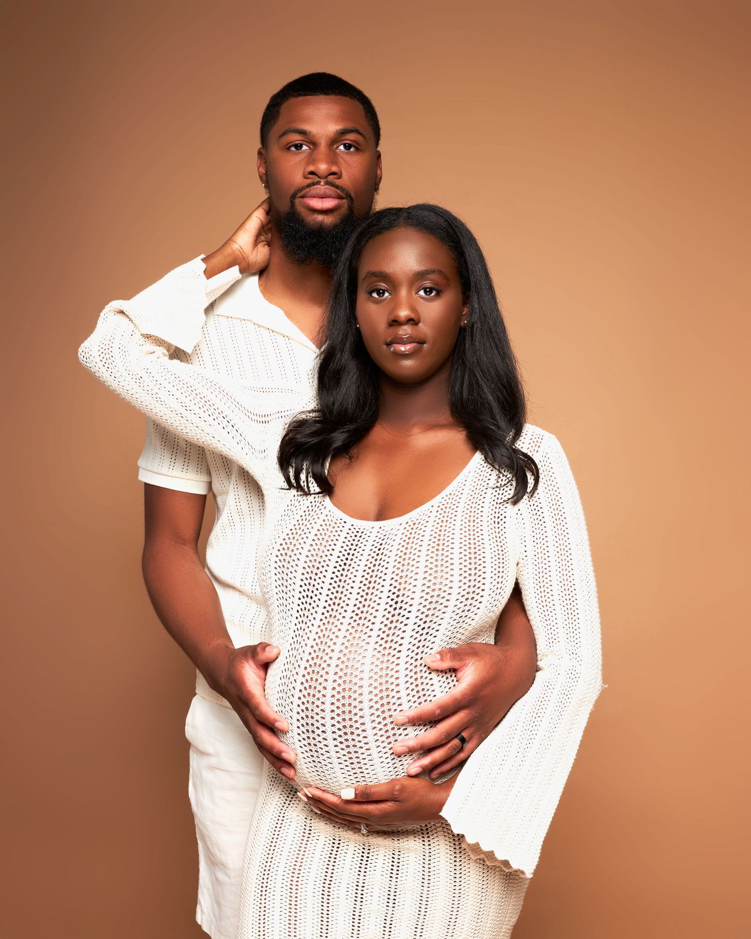 Maternity photo of couple in white knit outfits embracing against brown backdrop.