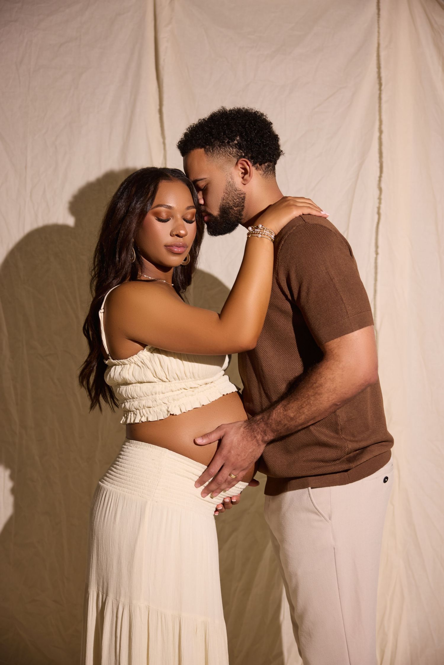 A couple shares an intimate moment, the man kissing the woman's forehead while cradling her baby bump.