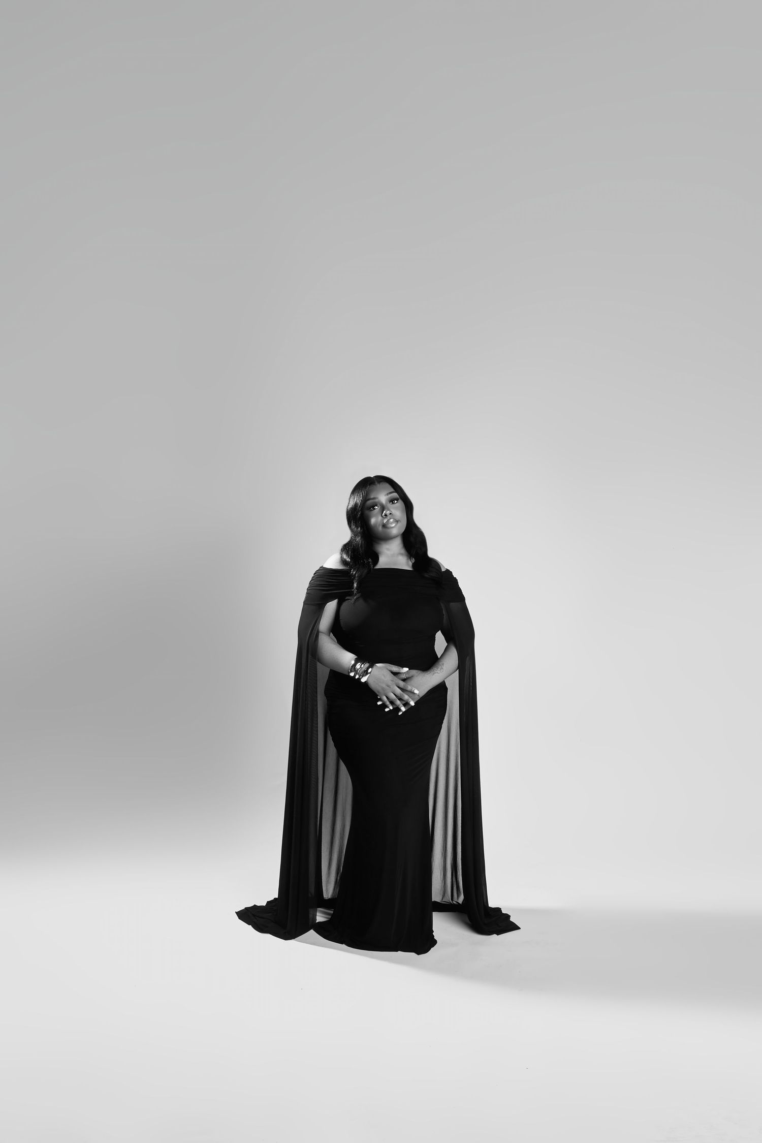 Woman in elegant black cape gown poses confidently in a black and white studio portrait.