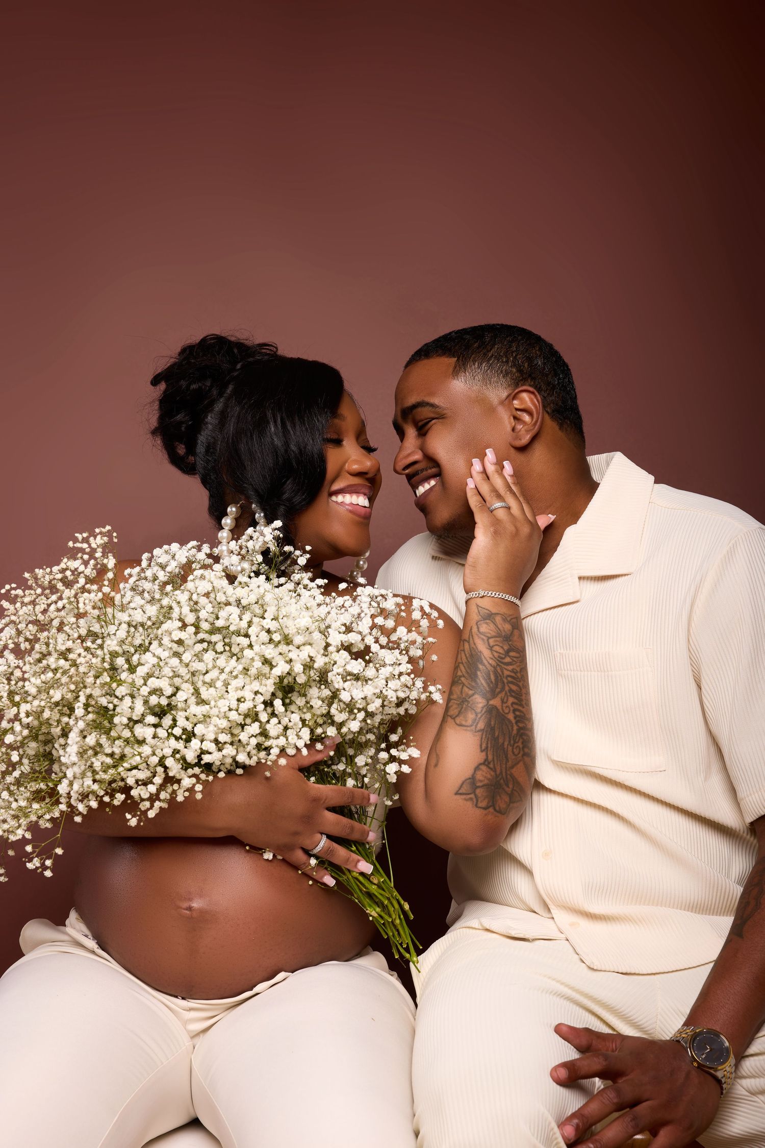 Intimate maternity portrait series with baby's breath flowers and cream-colored outfits against warm brown background.