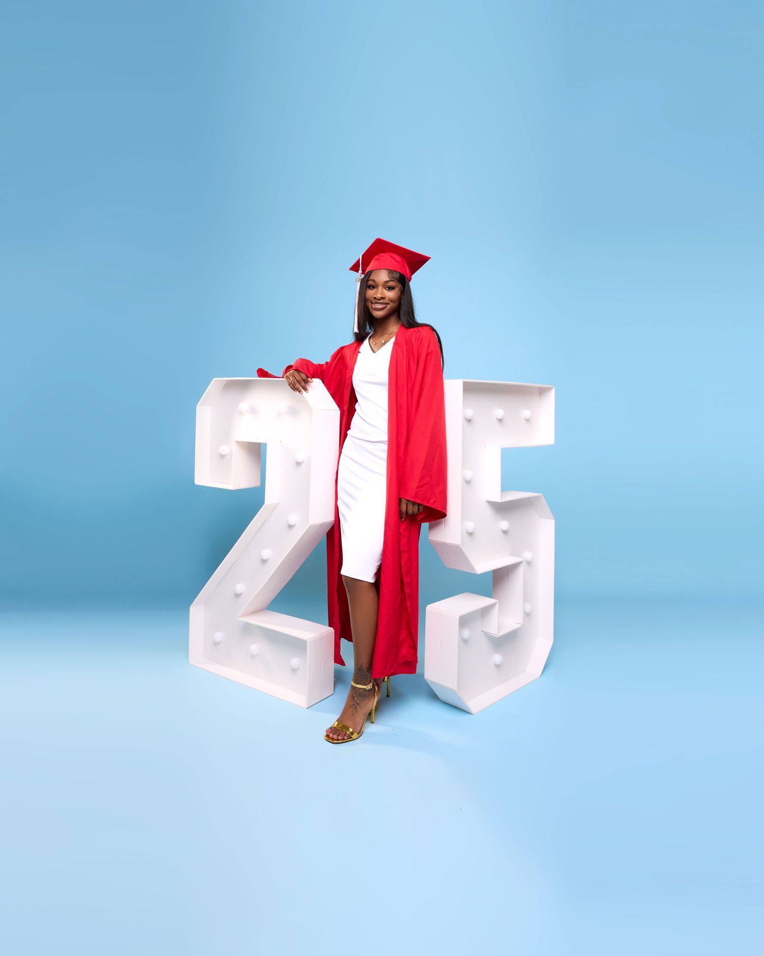 A graduate in red academic regalia poses with large white '25' numbers on blue background.