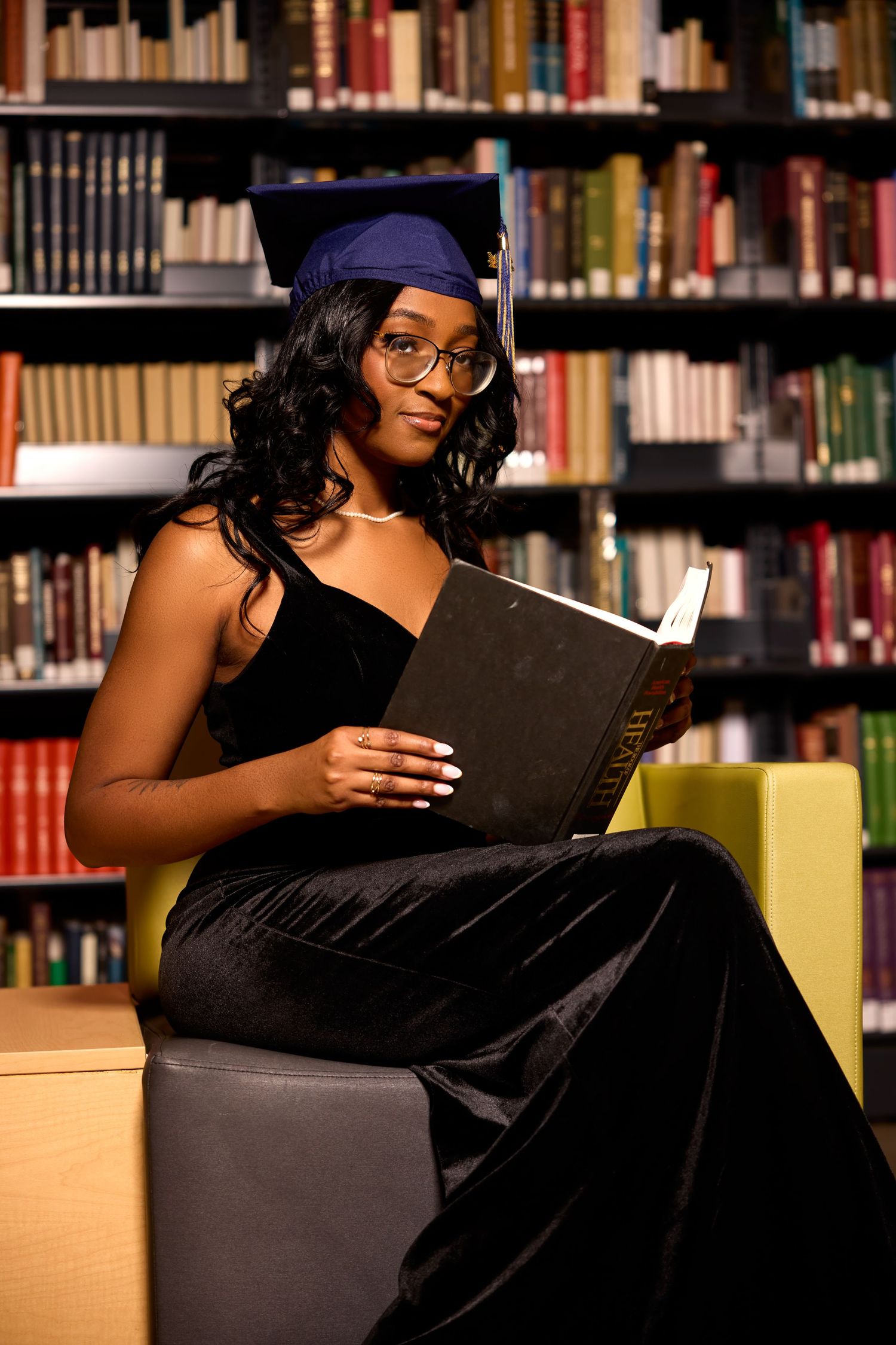 Graduate reading in library while seated on yellow chair surrounded by bookshelves.