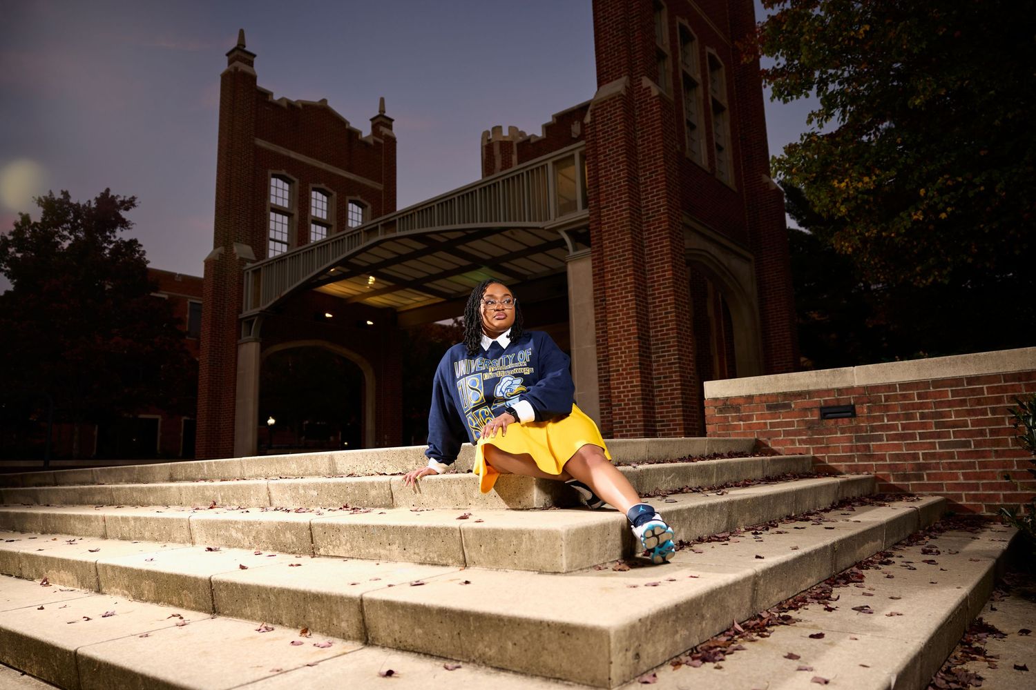 A student poses on concrete steps in front of a brick building with Gothic architecture at dusk.