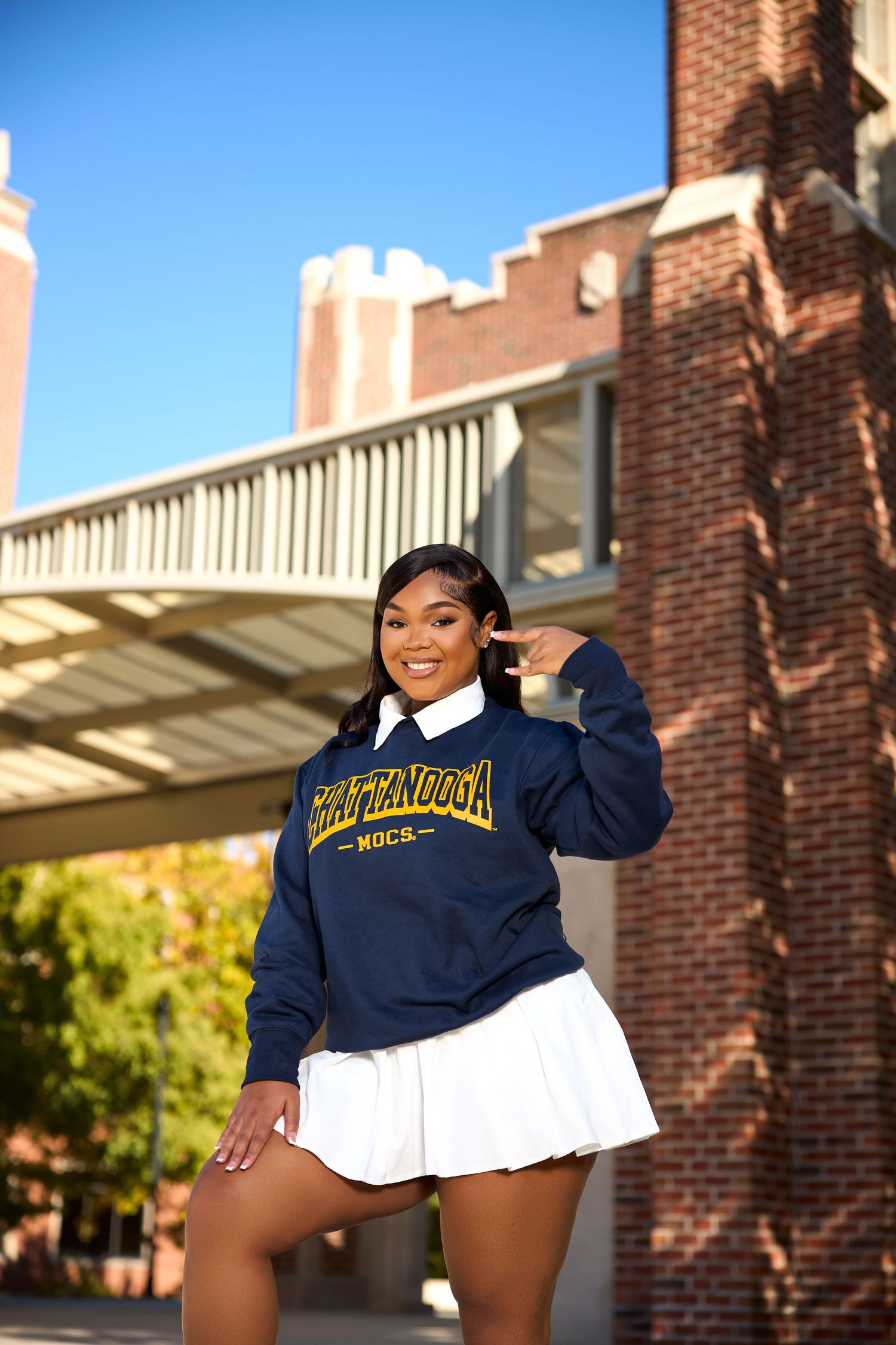 A student wearing a navy university sweatshirt and white skirt poses enthusiastically on campus steps.