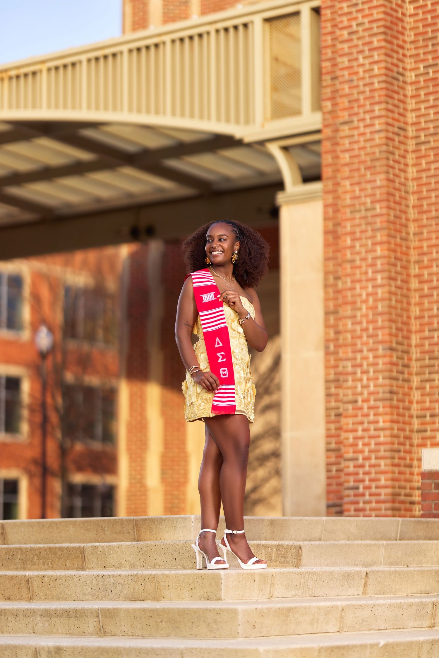 Young woman in gold dress holds Delta Sigma Theta stole on university steps, celebrating graduation achievement.
