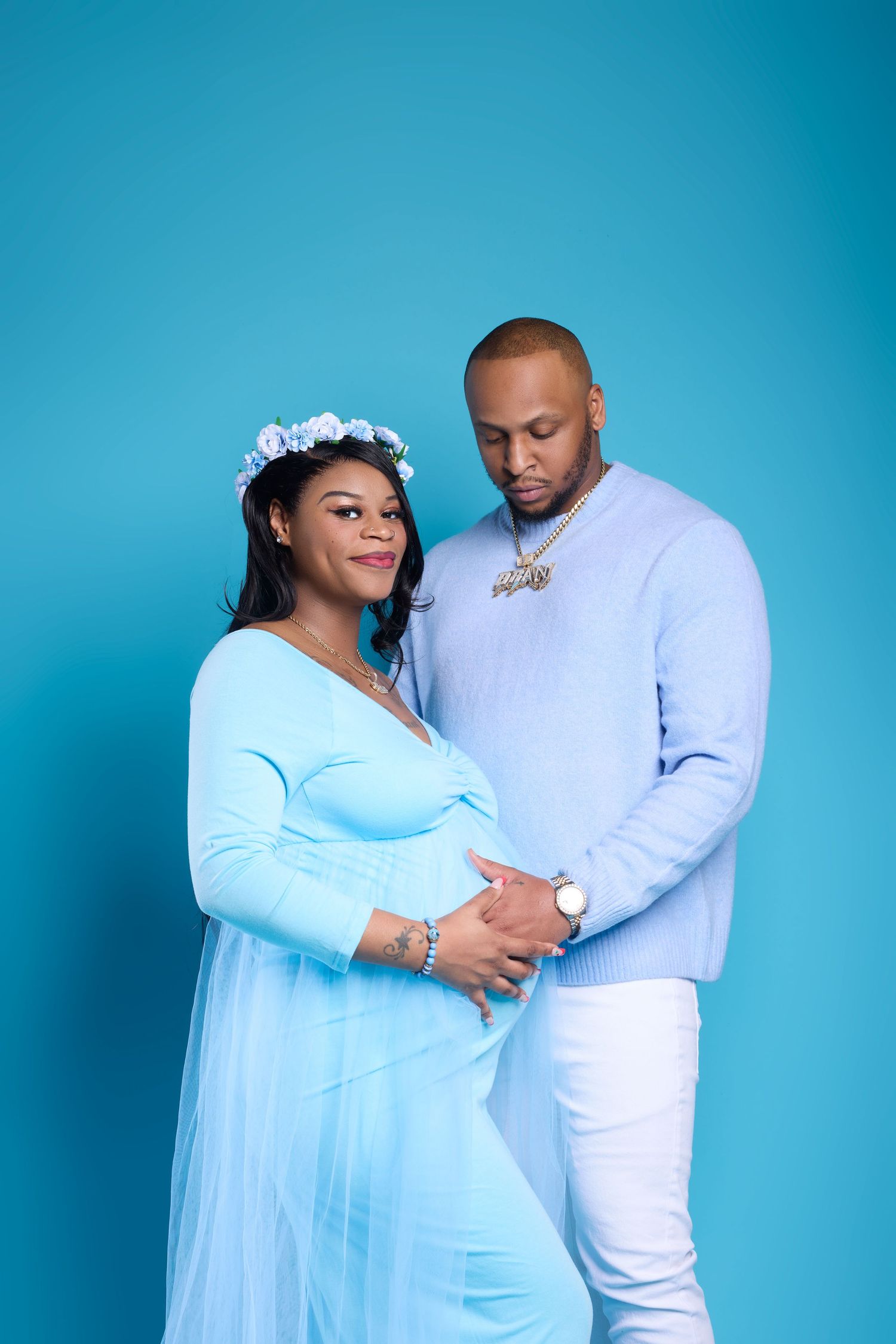 A pregnant woman in a blue dress and floral crown poses with a man in a light blue sweater against a blue background.