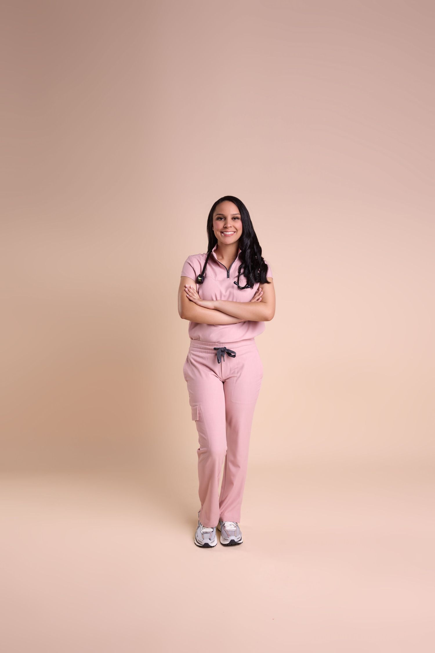 Smiling female nurse in pink scrubs with stethoscope, arms crossed, standing against a beige background.