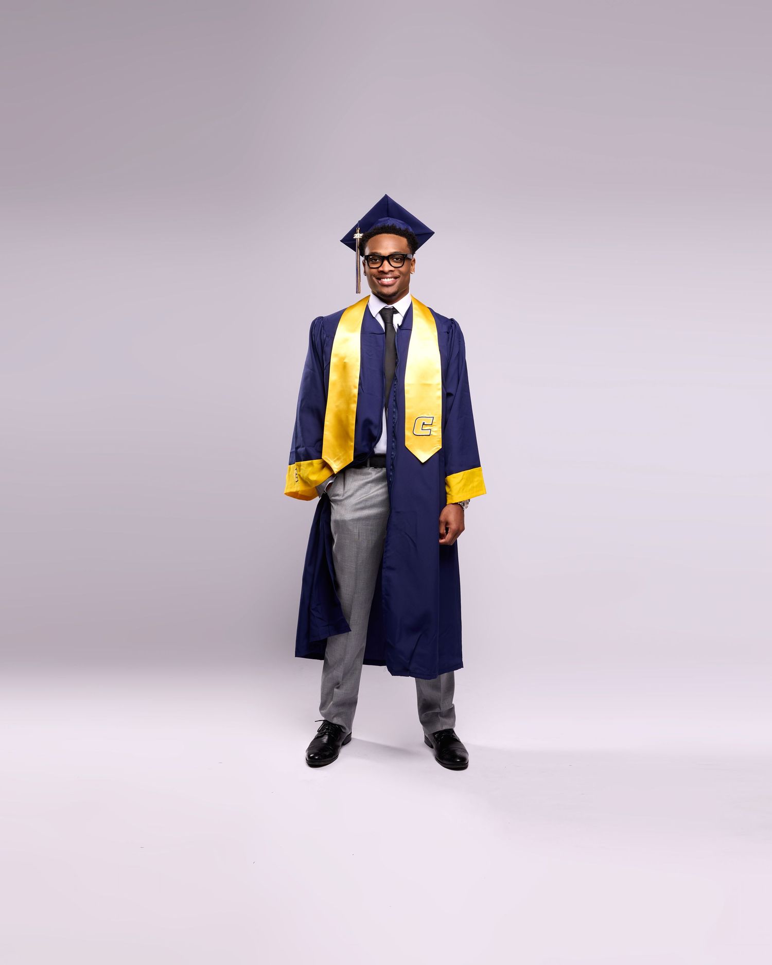 Graduate in blue cap and gown with gold stole poses against purple background.