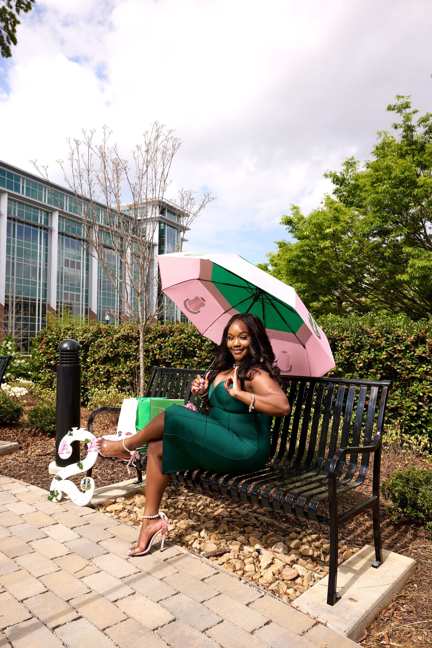 A graduate in green attire sits on a bench with pink and green umbrella near modern campus building.