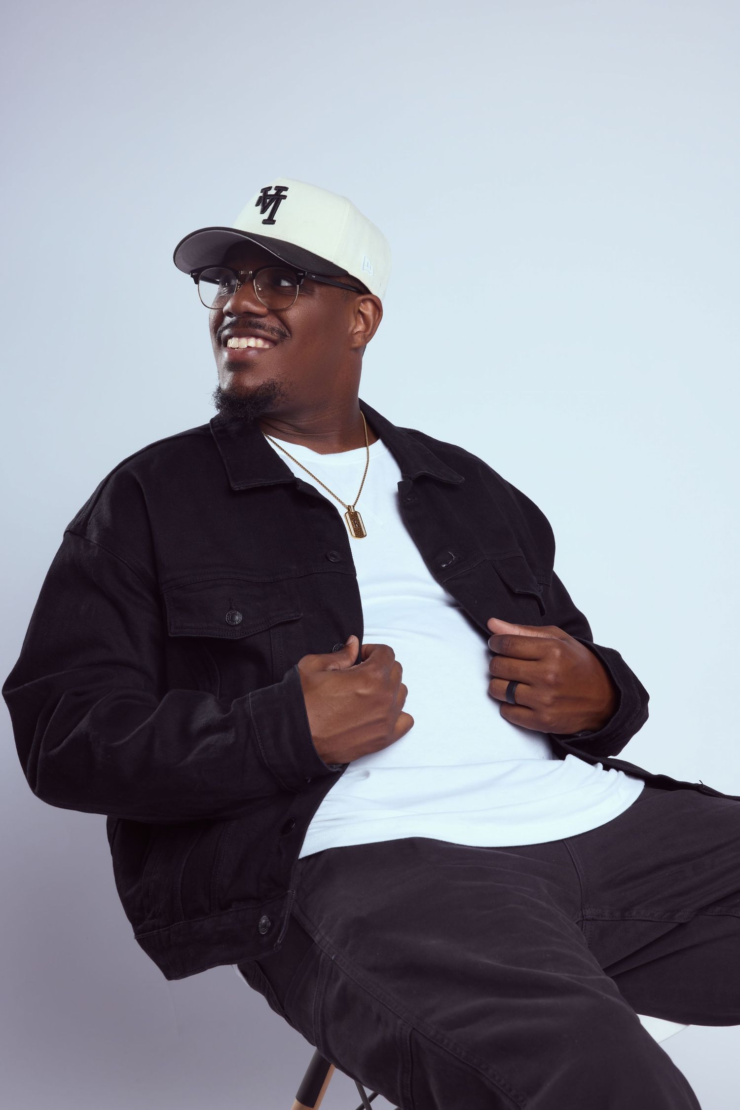 Smiling Black man in white cap, glasses, black jacket over white tee, gold chain, seated against light gray background.