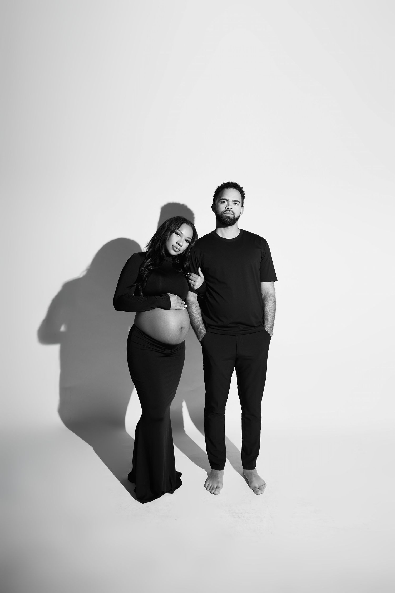 Black and white maternity photo of a pregnant woman in a gown with her partner in a studio setting.
