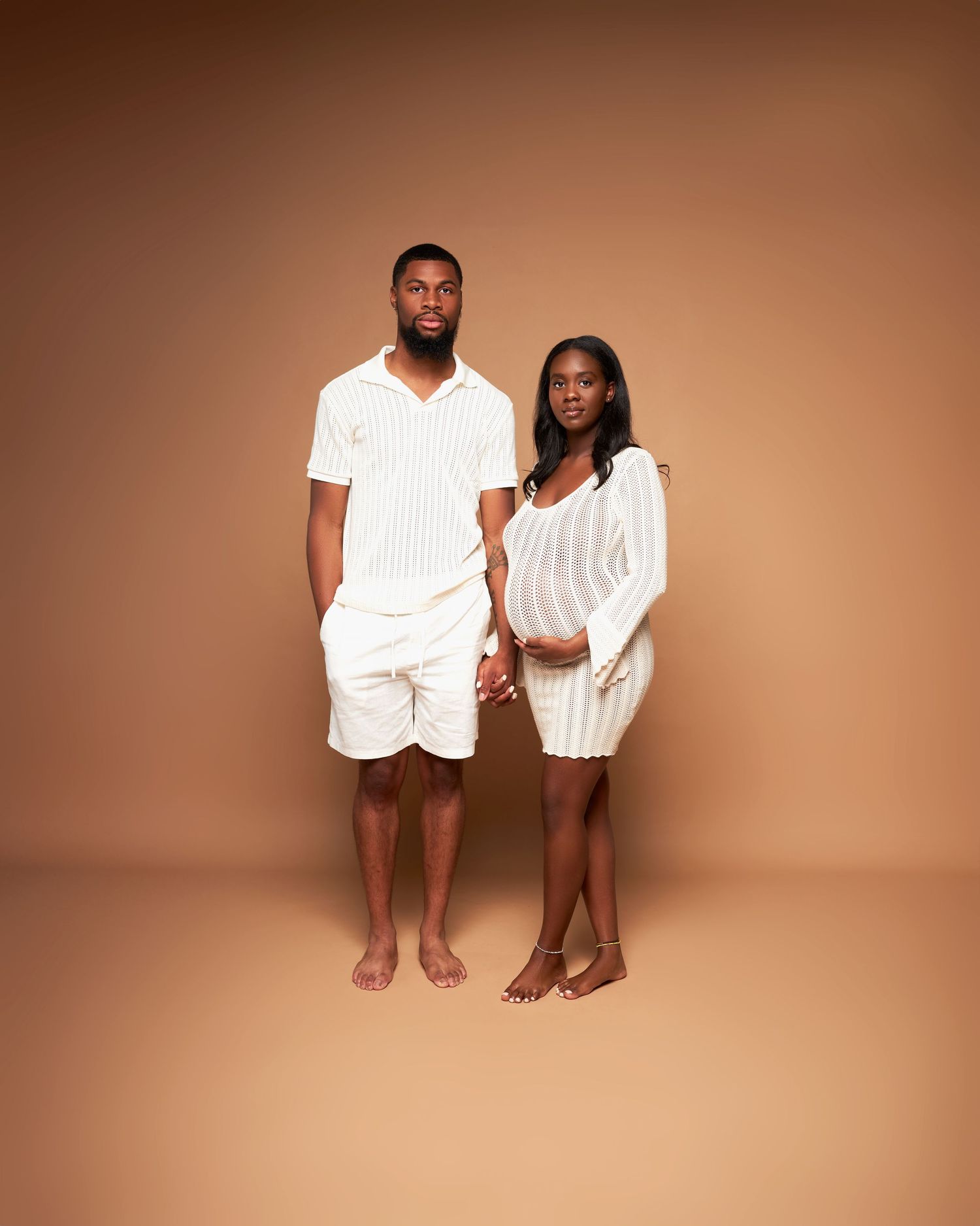 Couple wearing matching white outfits standing barefoot against warm brown background.