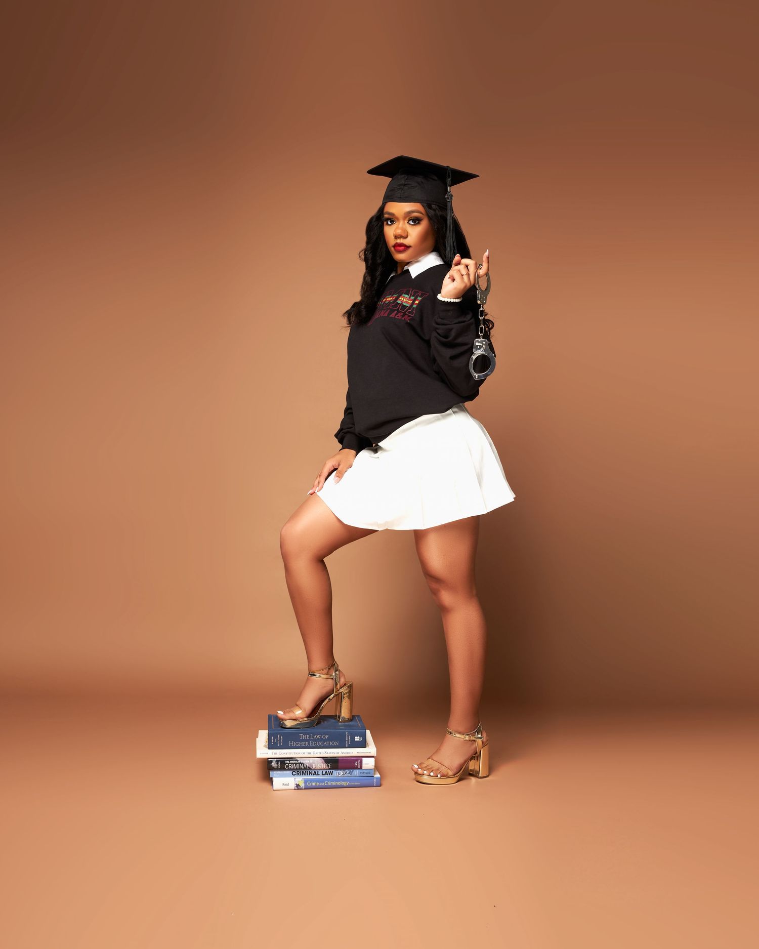 Graduate in black cap and gown poses with books in artistic studio photoshoot against brown backdrop.