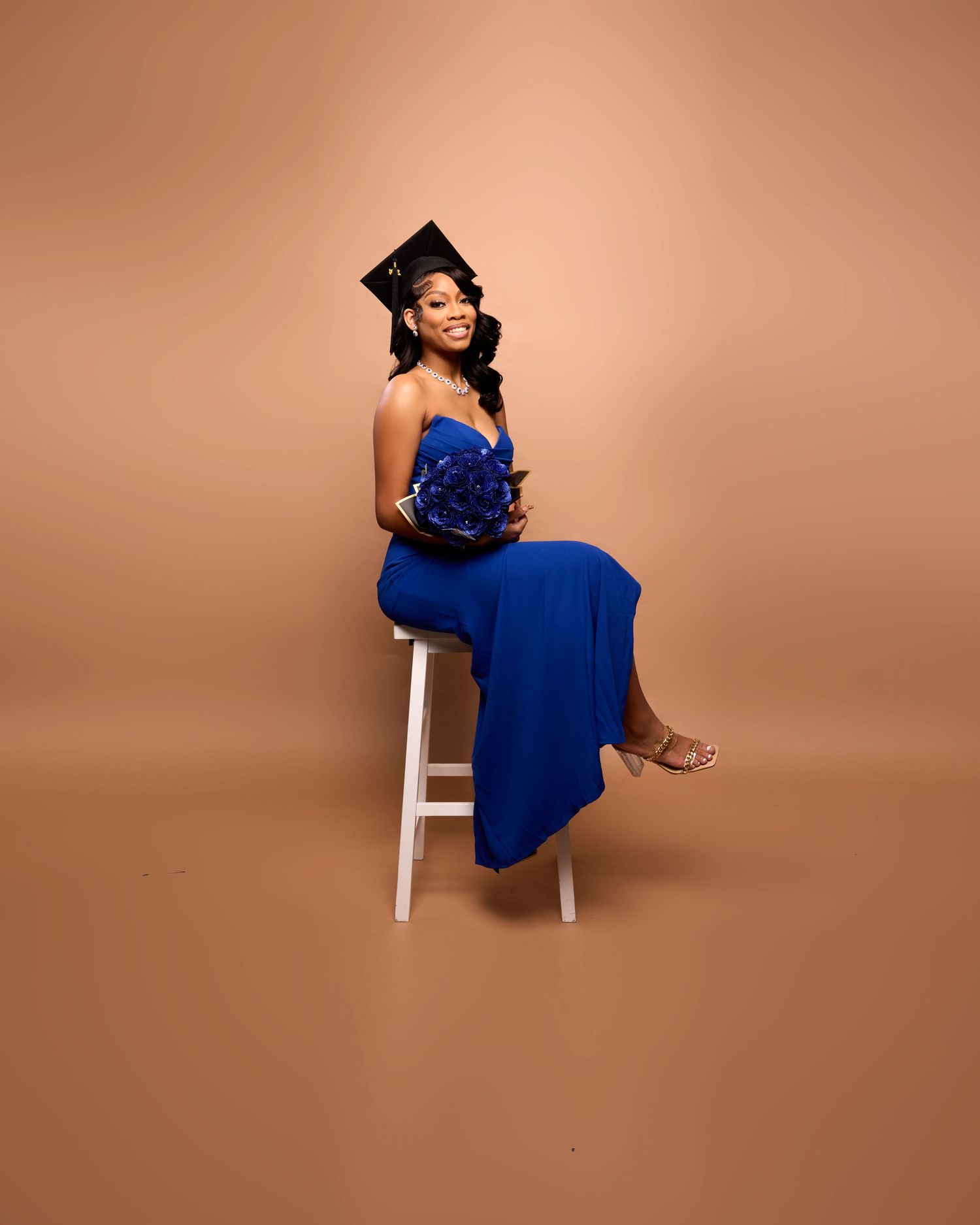 Person in blue formal dress poses elegantly on white chair against peachy background.