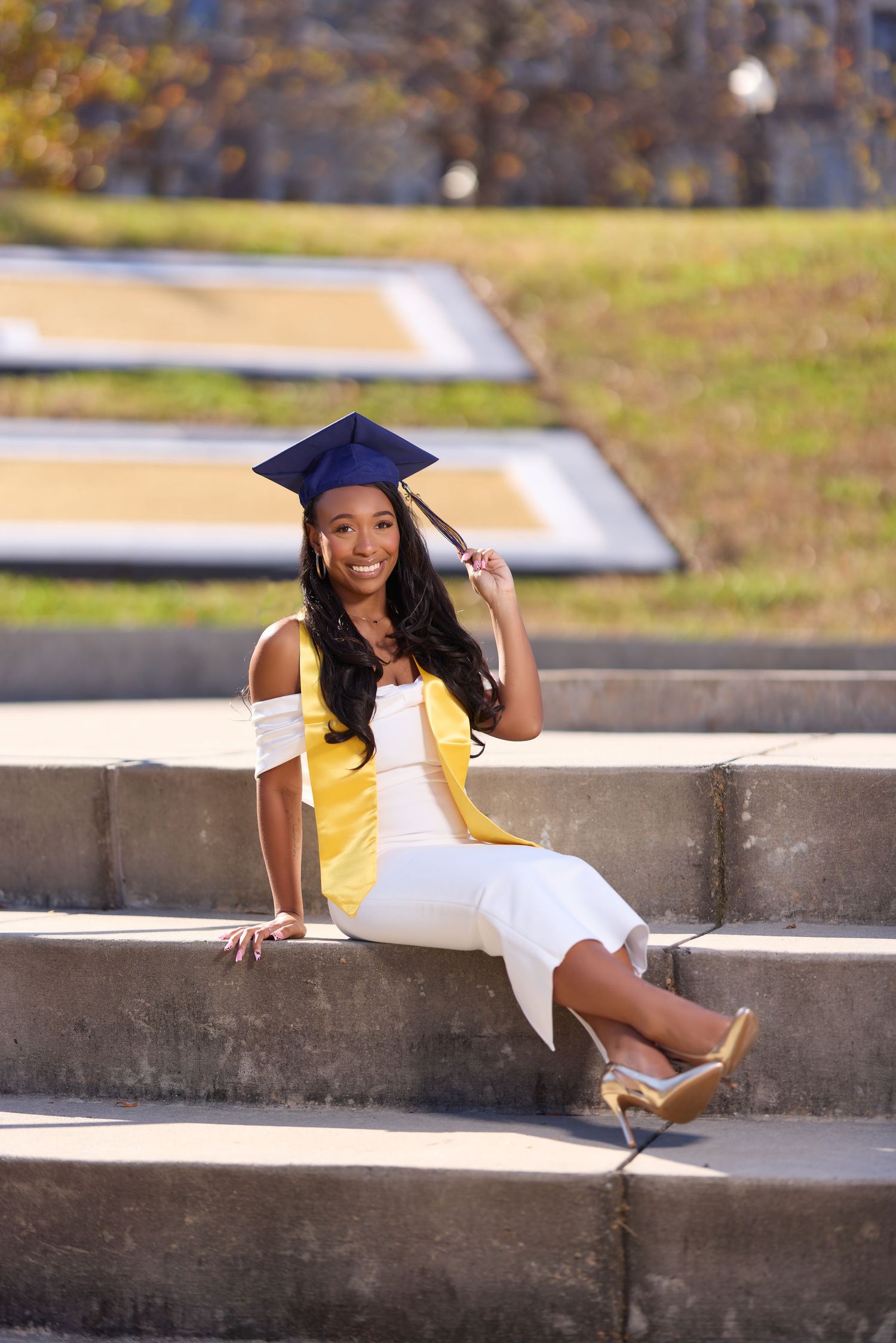 Graduate in yellow top and cap poses playfully on concrete steps outdoors.