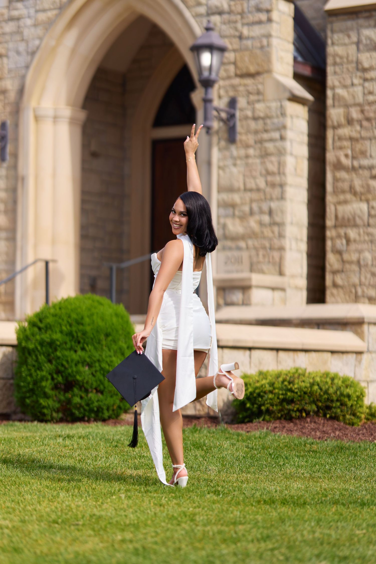 Smiling graduate in white dress holding cap, celebrating outdoors near stone building on green lawn.