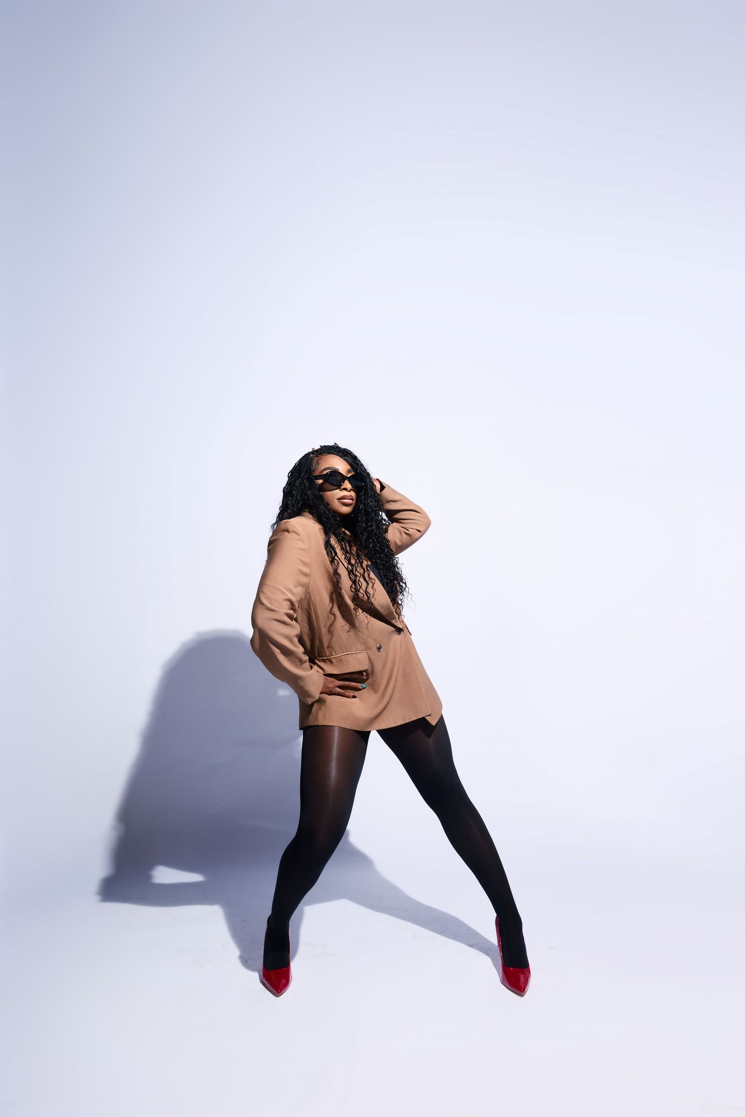 Stylish woman in beige blazer, black tights, and red heels posing confidently on white background.