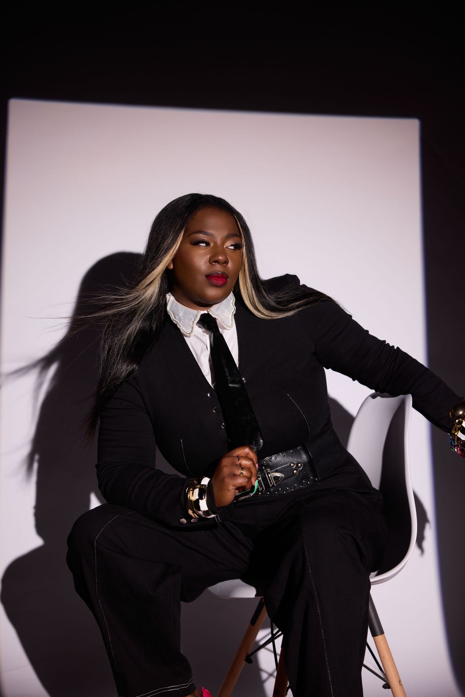 A confident woman in a black suit with white shirt sits on a chair, featuring blonde-streaked hair against a dramatic backdrop.