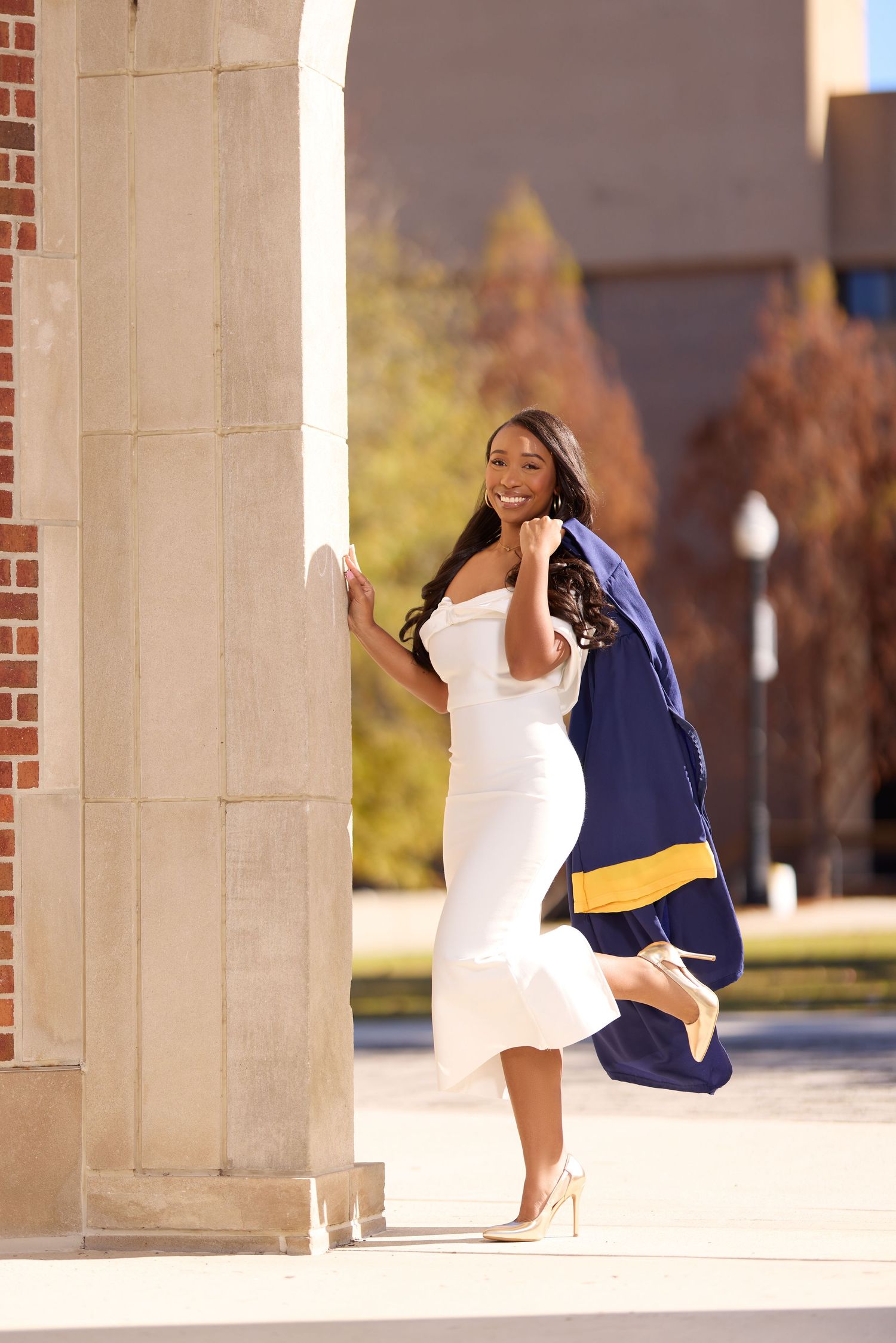 Graduate in white dress and navy gown poses between architectural columns.