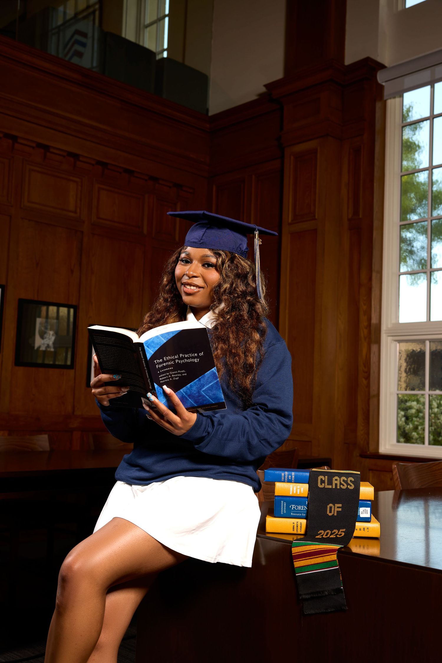 Graduate reads from textbooks while posing in classic university library interior.