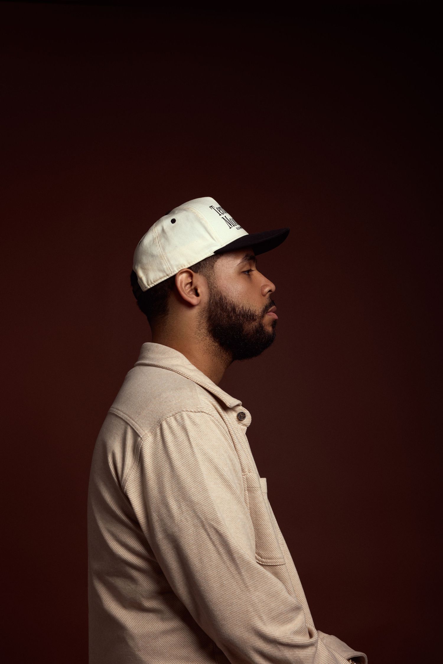 Profile portraits of person in white cap and beige jacket against dark brown backdrop.