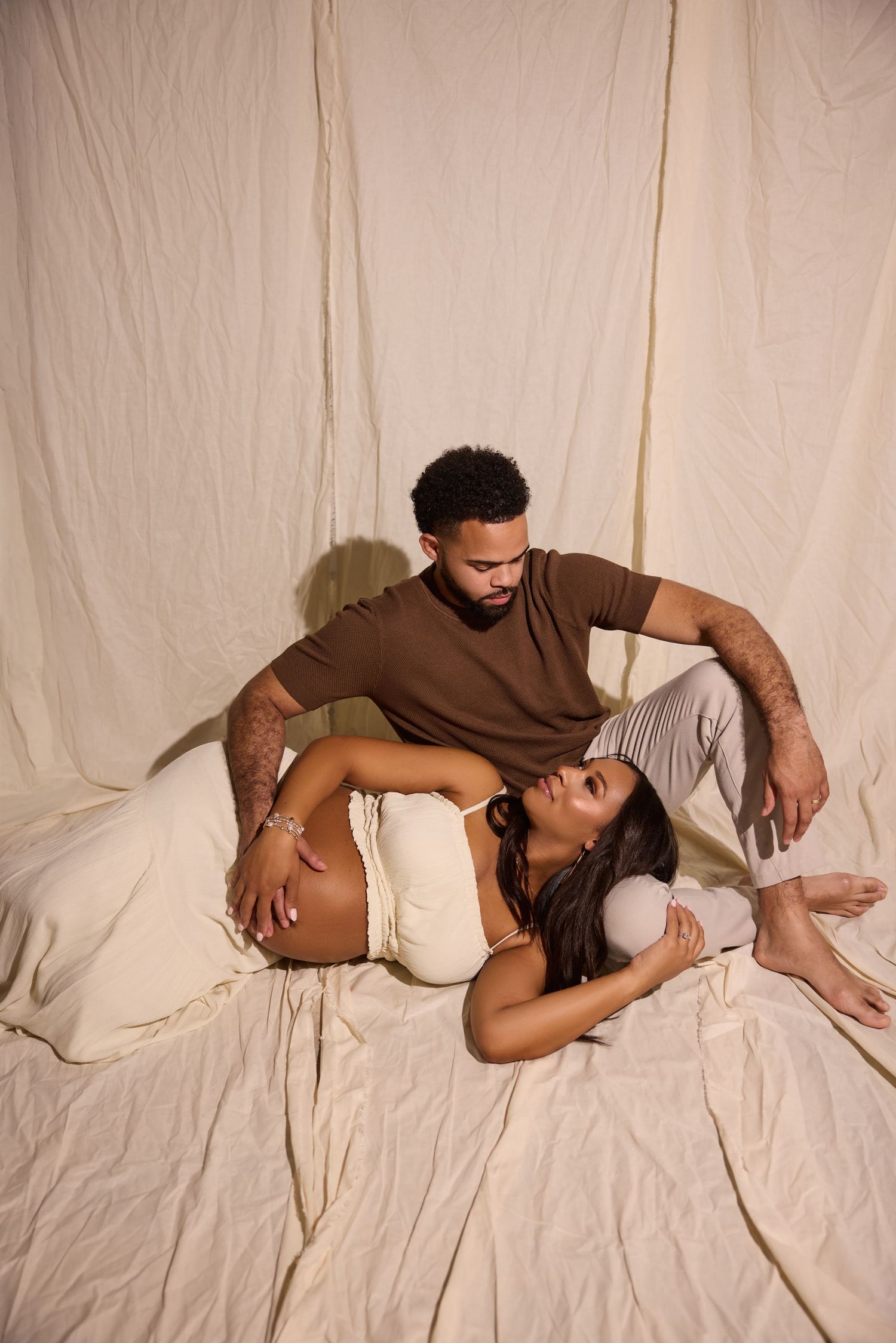 A couple relaxes on a bed, woman lying down smiling up at man seated beside her, both in neutral tones against cream fabric backdrop.