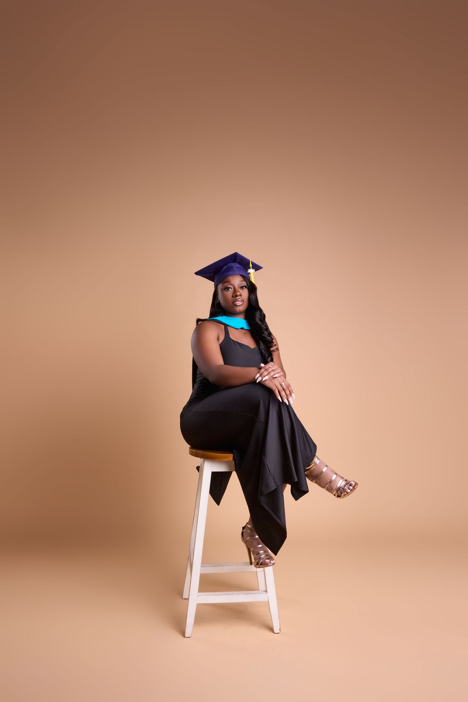 Young Black woman in graduation cap and gown seated on white stool against beige background, celebrating academic achievement.