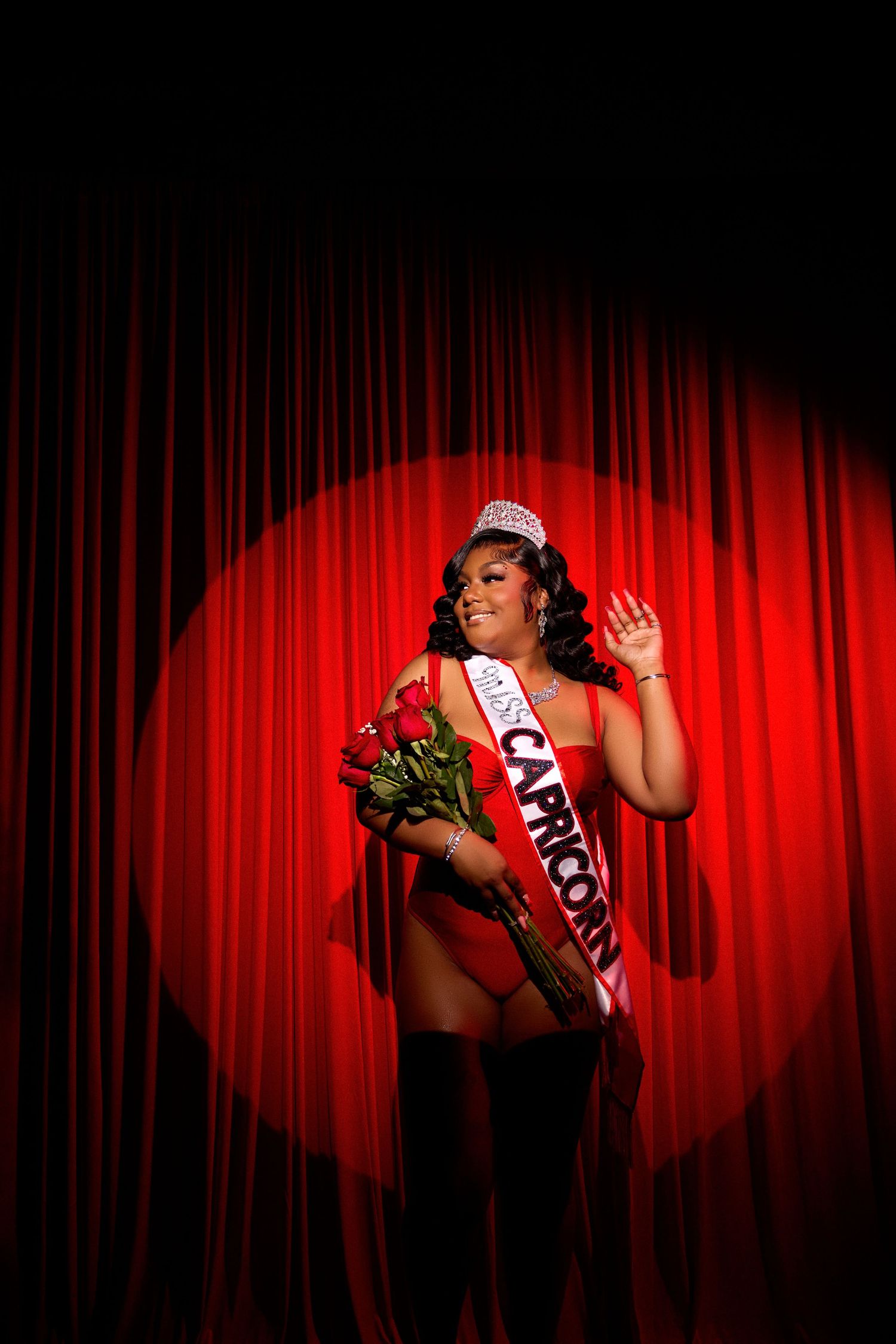Pageant contestant poses playfully in front of red curtain wearing sash and crown.