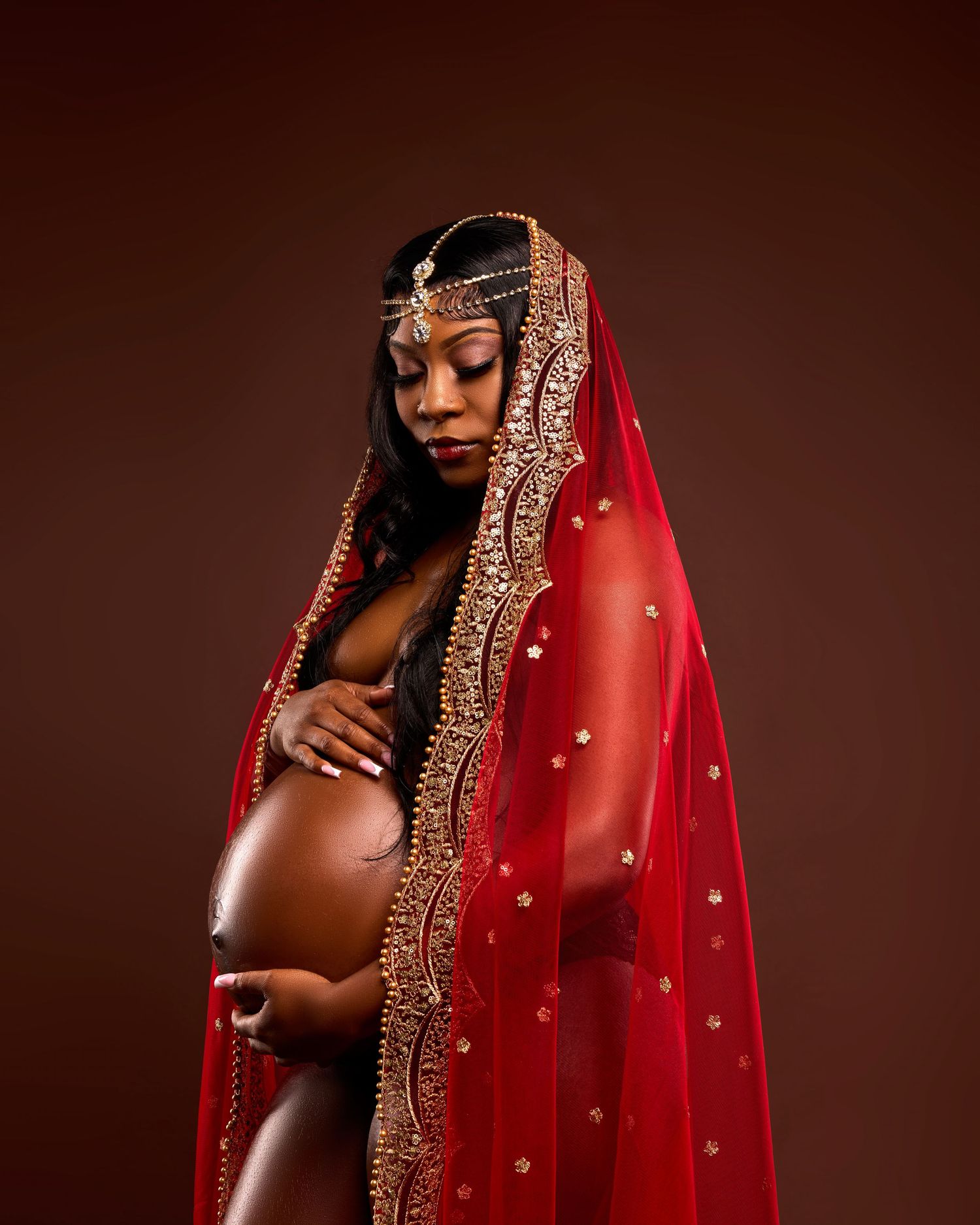 Artistic maternity portrait with woman draped in ornate red and gold traditional veil.