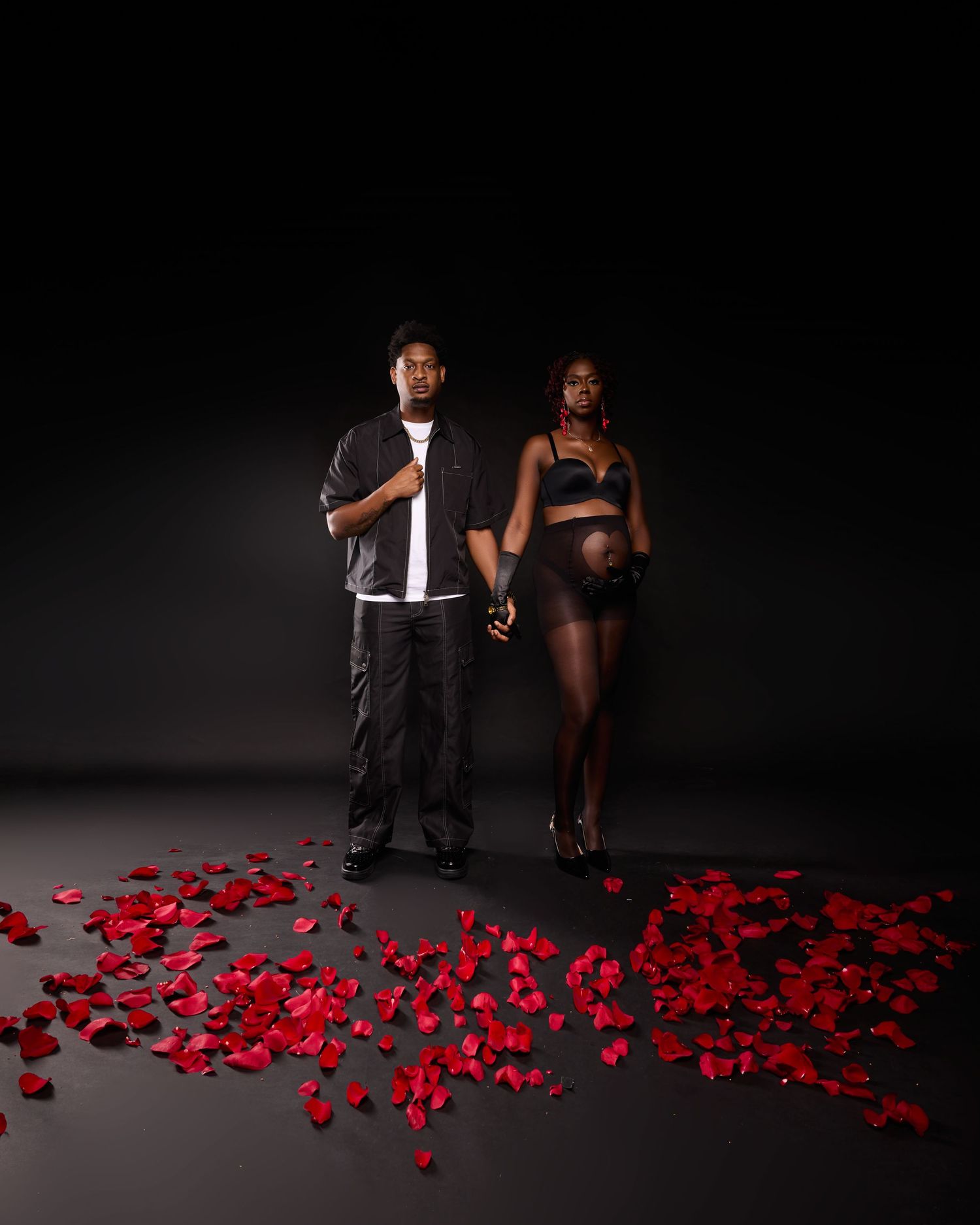 Moody maternity photos with couple posed among scattered red rose petals against dark background.
