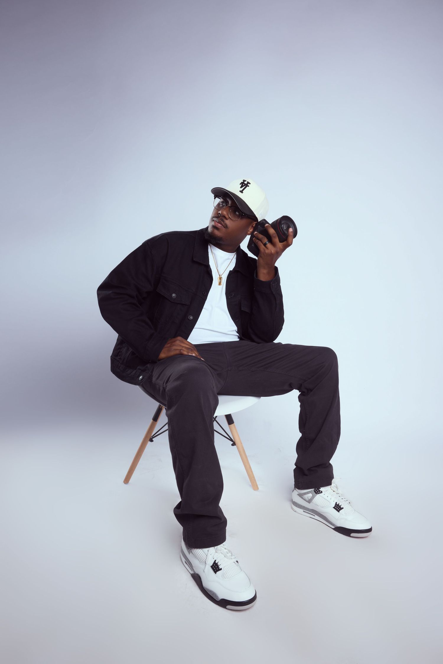 A young Black man sits on a wooden chair, wearing a black jacket, dark pants, white sneakers, and a white cap, holding a camera.