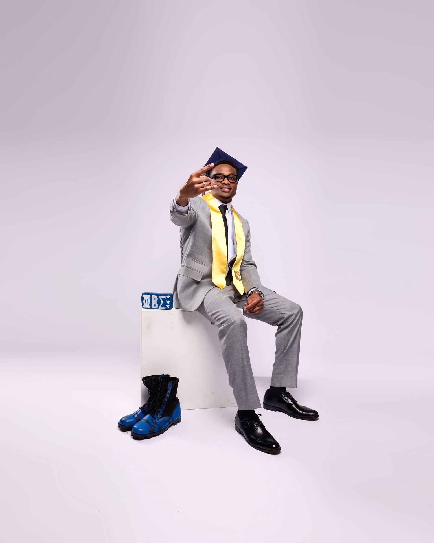 Graduate in formal attire and academic regalia poses on purple studio backdrop.