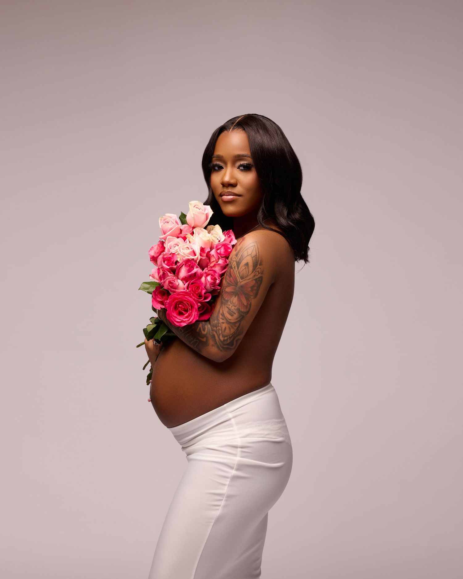 Artistic maternity photo of expectant mother holding pink roses against grey background.