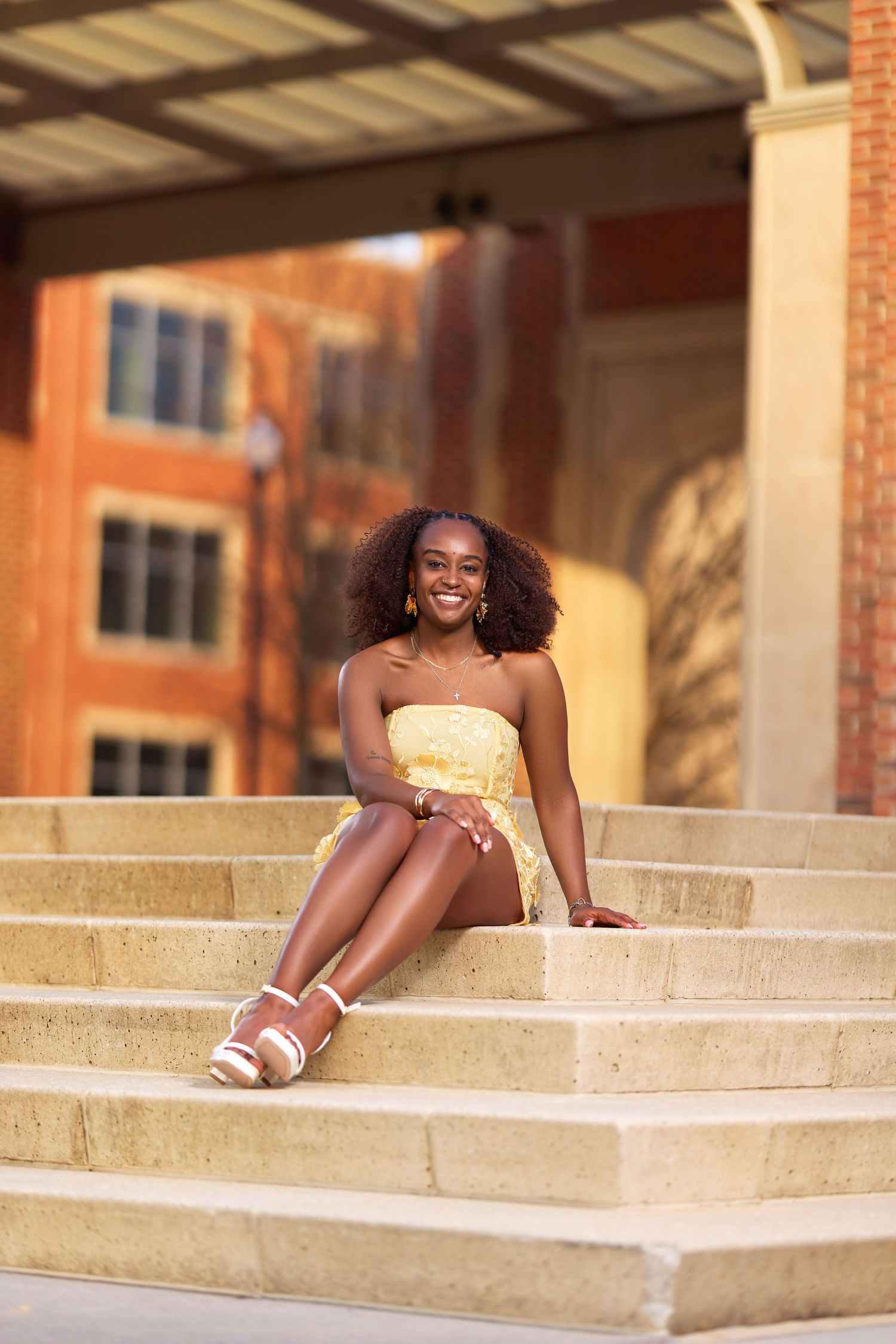 Young Black woman in yellow dress sitting on stone steps, smiling confidently outdoors near brick building.