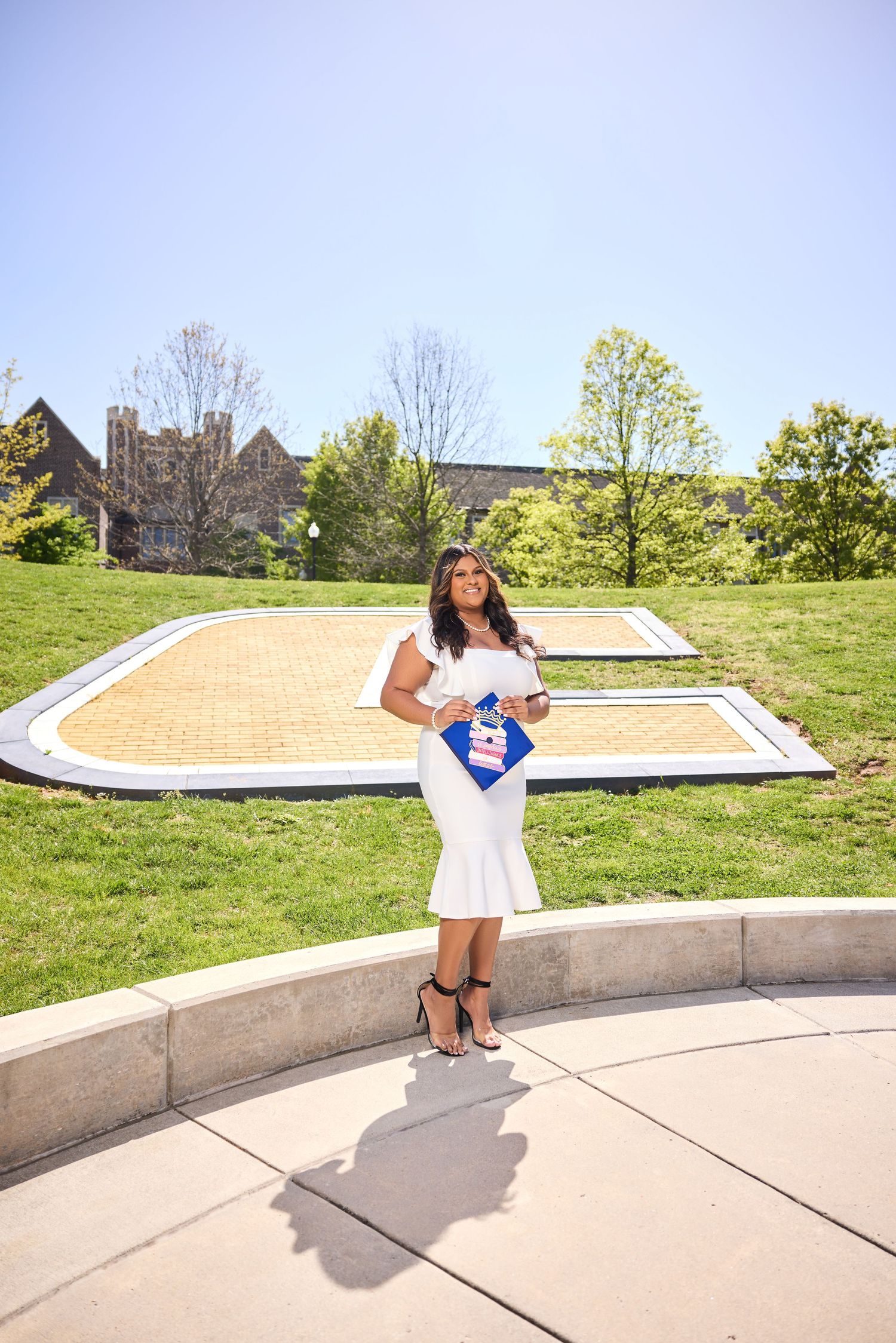 Graduate poses in white dress and blue sash on concrete steps with grassy hill and buildings in background.
