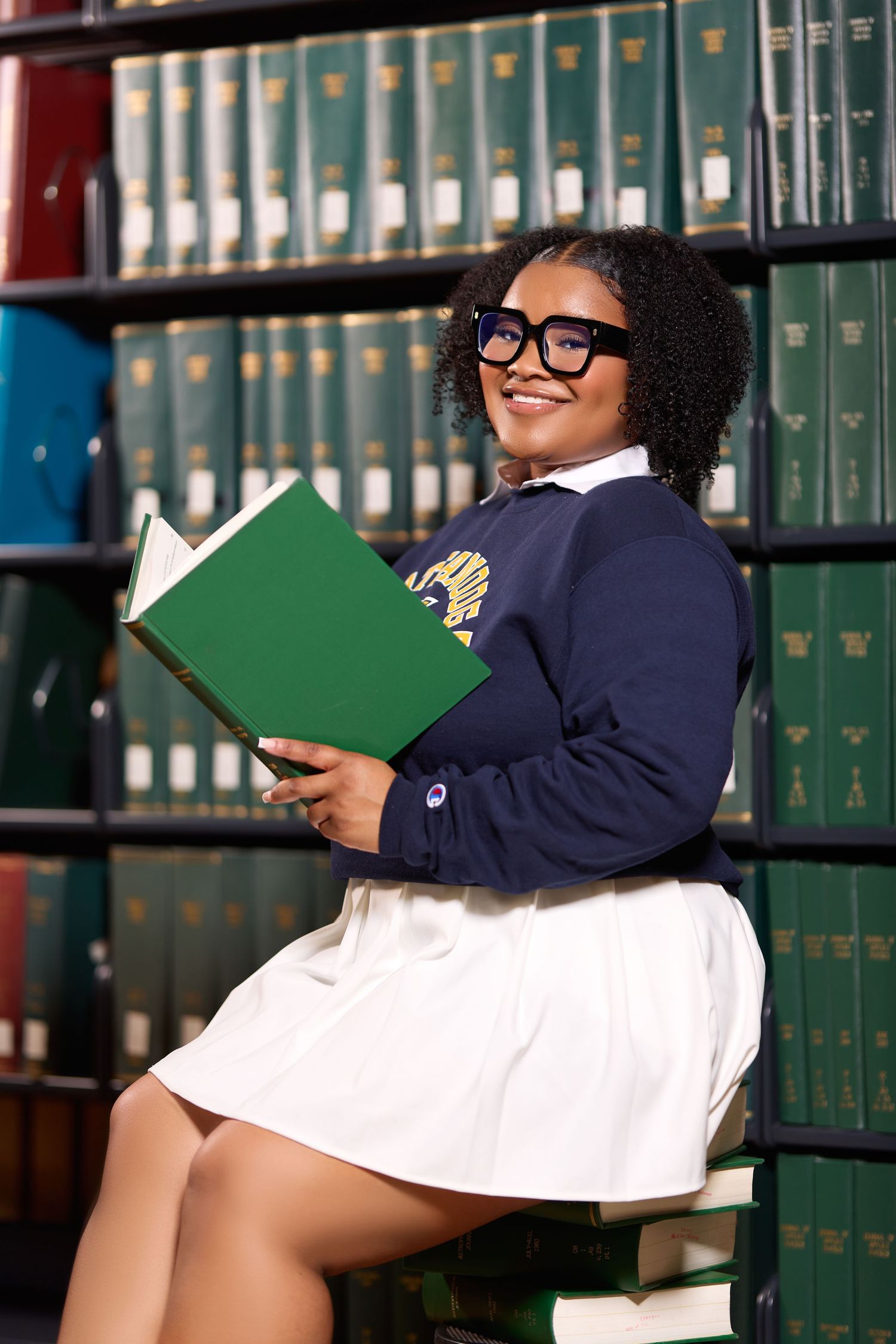 Student in glasses poses with book in front of library reference shelves.