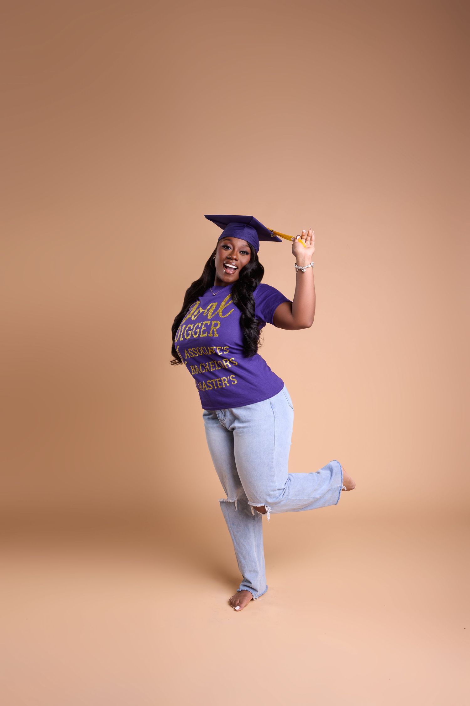 A joyful woman in a purple graduation cap and t-shirt with ripped jeans poses playfully against a beige background.