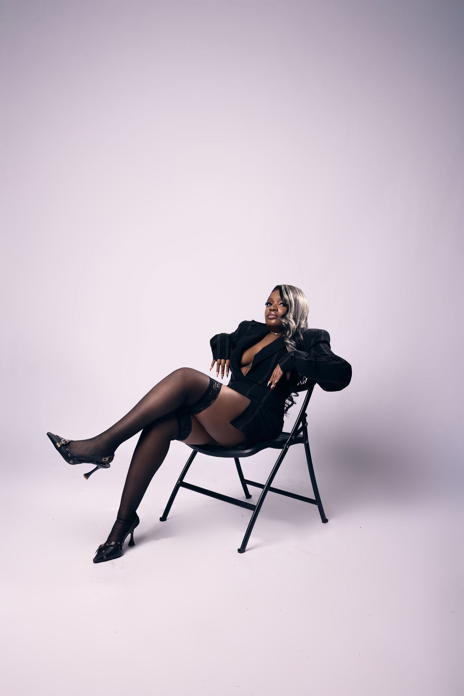Artistic pose on folding chair with black outfit against white studio backdrop.