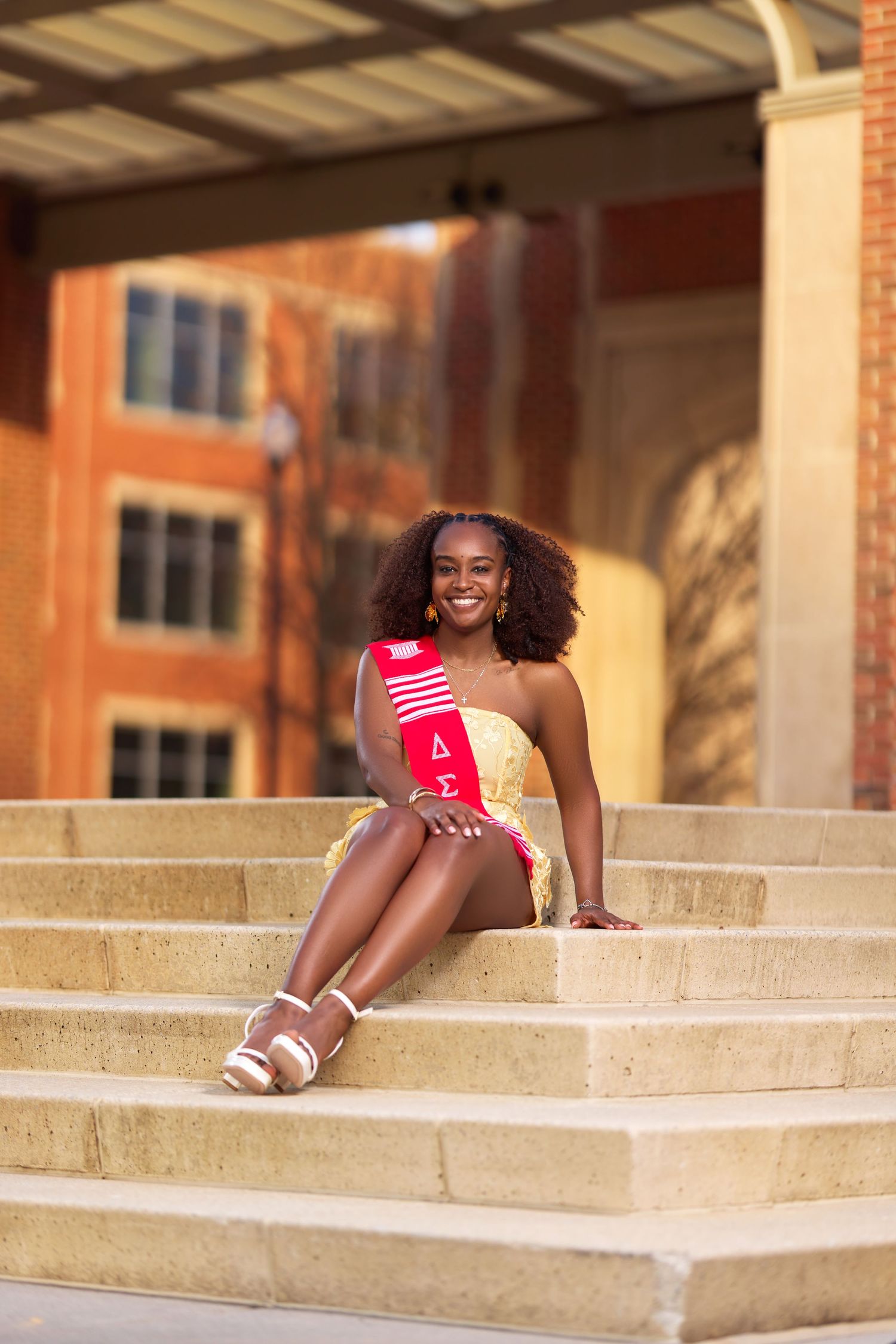 Smiling young woman in yellow dress wearing Delta Sigma Theta sash sitting on stone steps outside brick building.