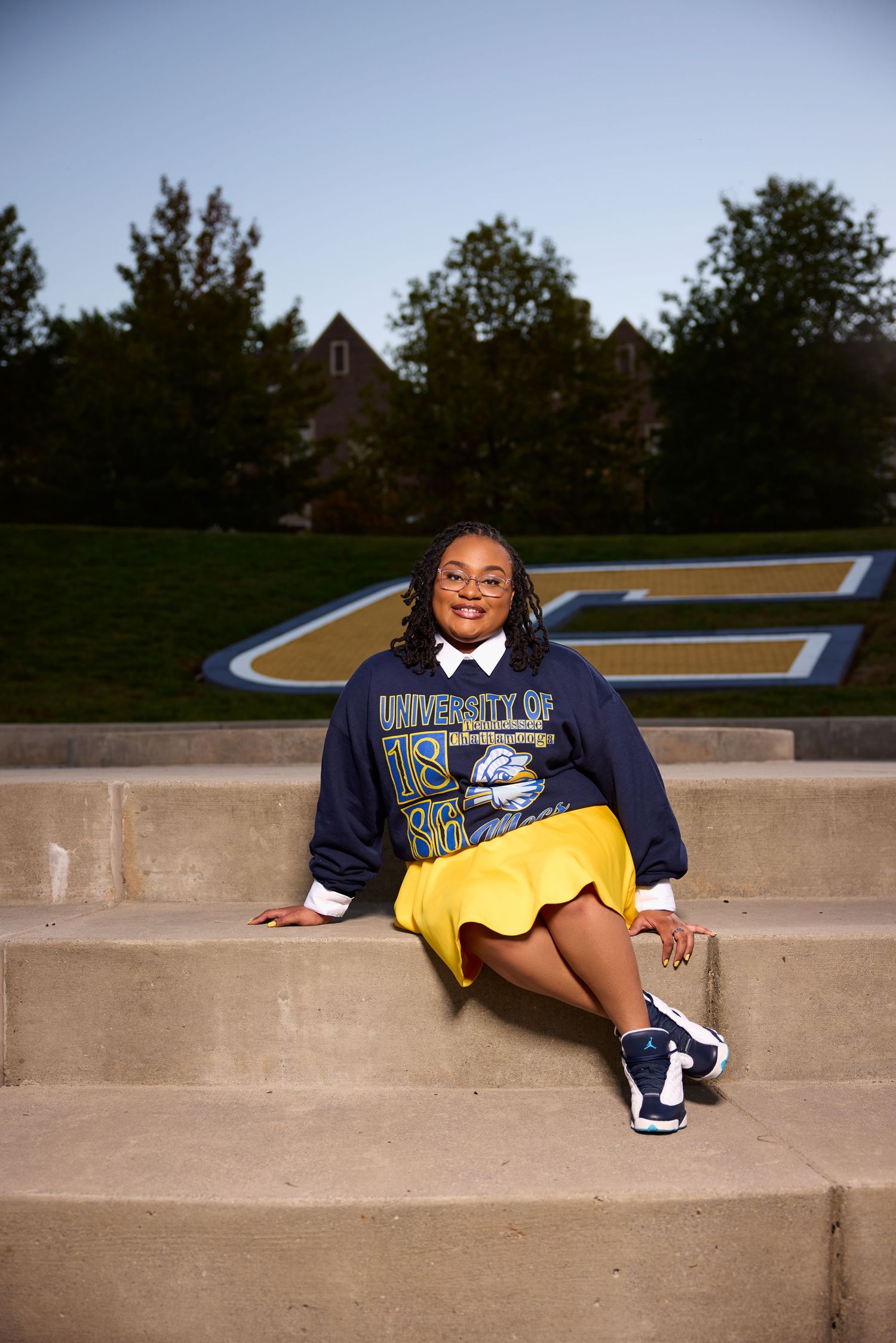 Graduate in navy and yellow attire poses on concrete amphitheater steps outdoors.