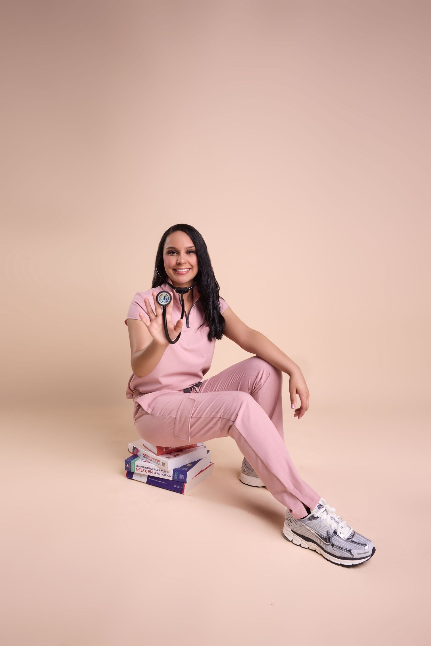 A smiling female doctor in pink scrubs sits on medical books, holding a stethoscope against a beige background.